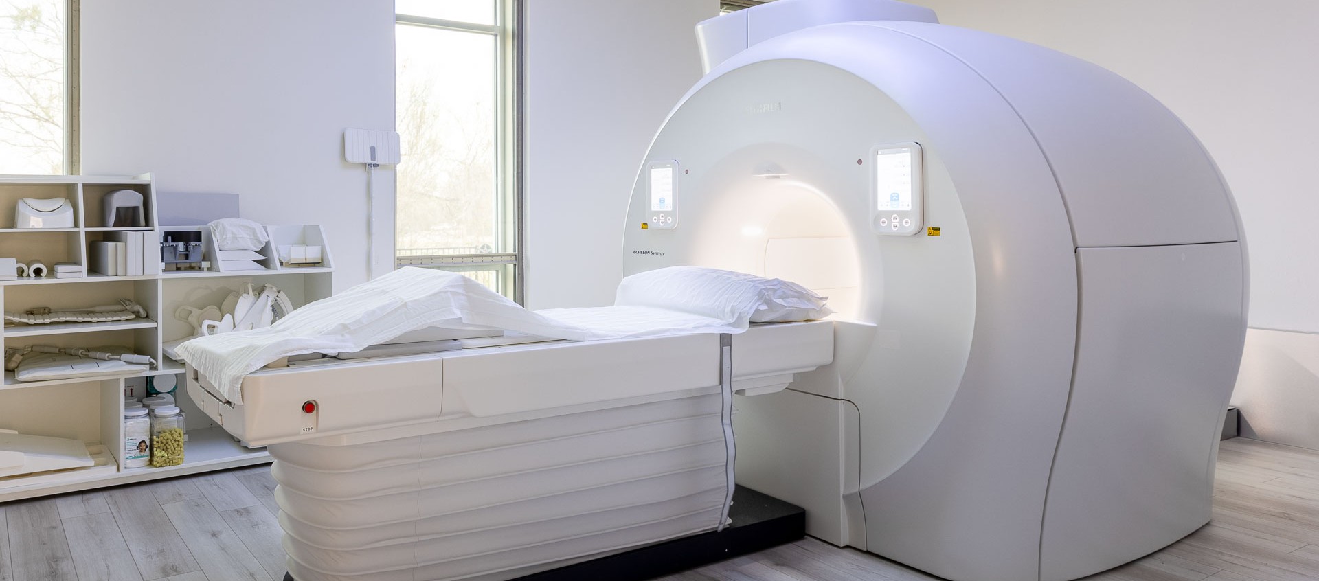 The 1.5T Fujifilm Synergy CT machine with a white padded patient table and soft overhead lighting in a clean medical room.