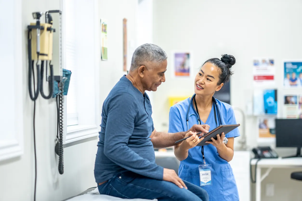 A senior gentleman of Hispanic decent, sits up on an exam table during a routine check-up at the doctors. A nurse, who is wearing blue scrubs, is standing in front of the gentleman as she reviews some recent test results with the patient on a tablet.