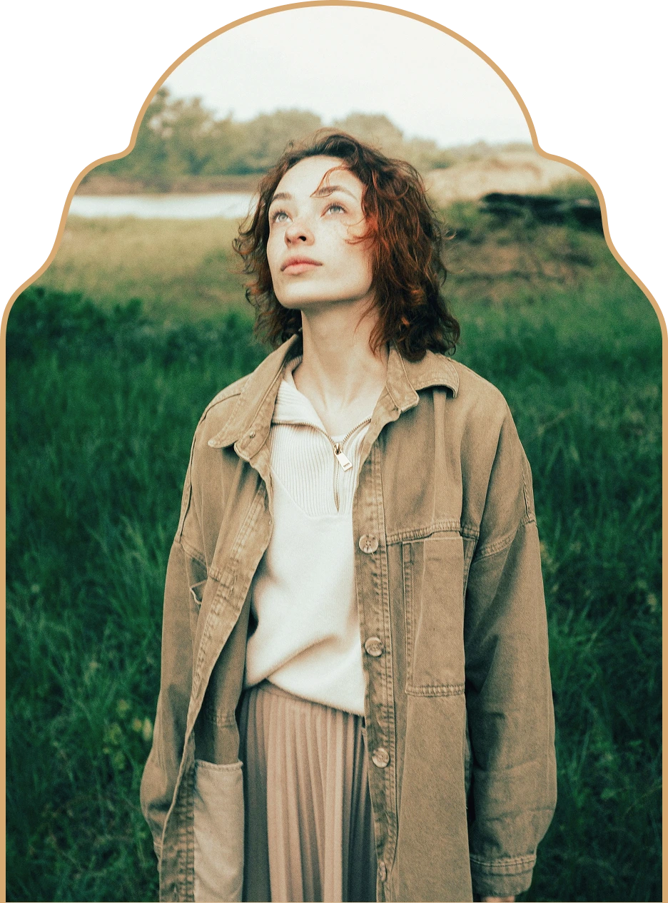 Young woman with curly hair wearing a beige jacket and light sweater looking up outdoors with green grass and trees in the background.