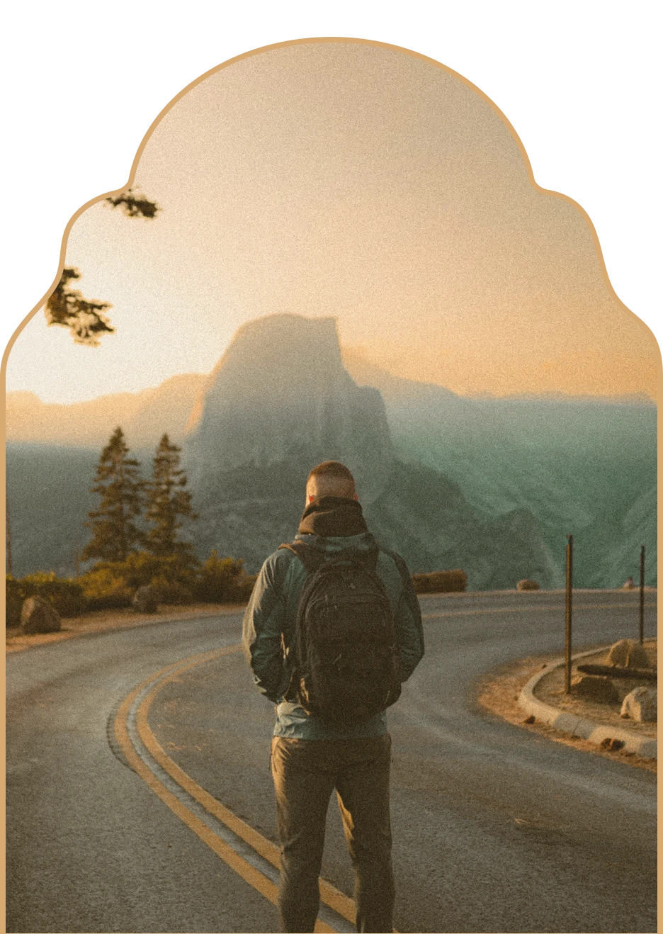 Man wearing a backpack standing on a winding road overlooking a mountain at sunset.