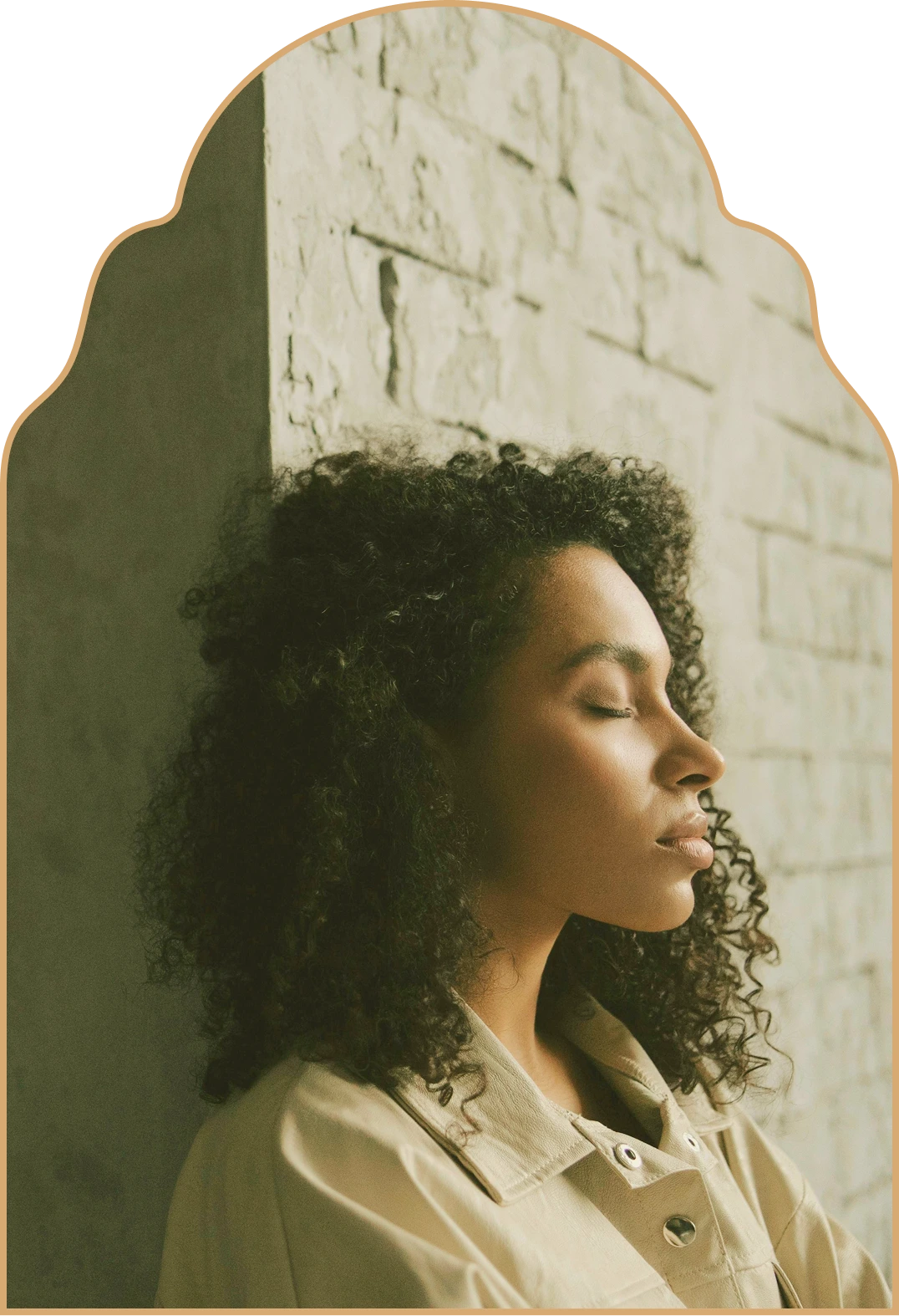 Side profile of a woman with curly hair and closed eyes leaning against a textured stone wall.