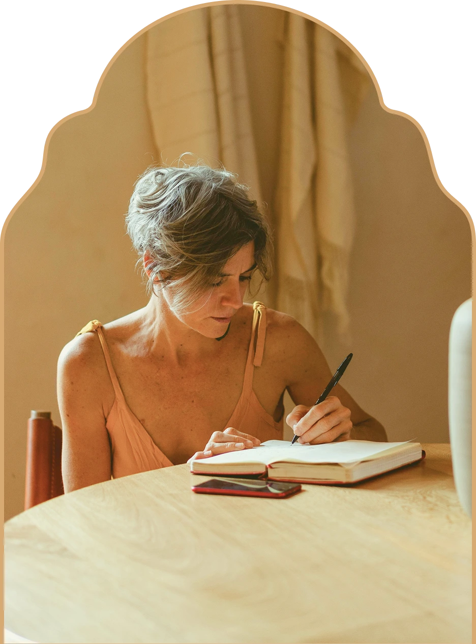 Woman with short hair writing in an open notebook at a wooden table with a smartphone next to it.