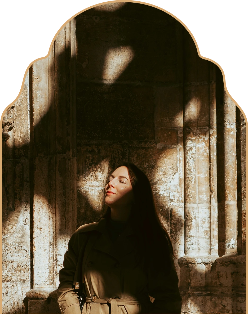 Woman standing with eyes closed, basking in sunlight against an old textured stone wall.