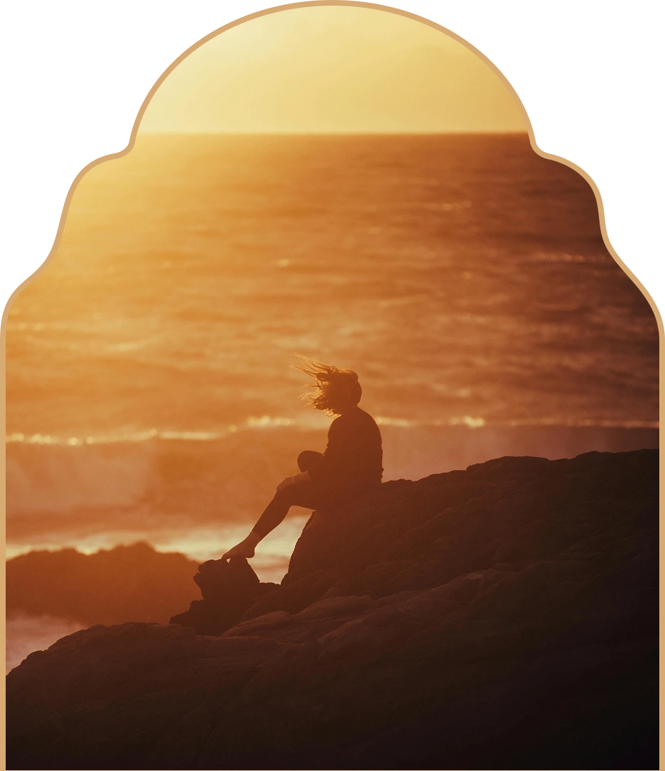 Silhouette of a person sitting on rocks by the ocean at sunset with wind blowing their hair.