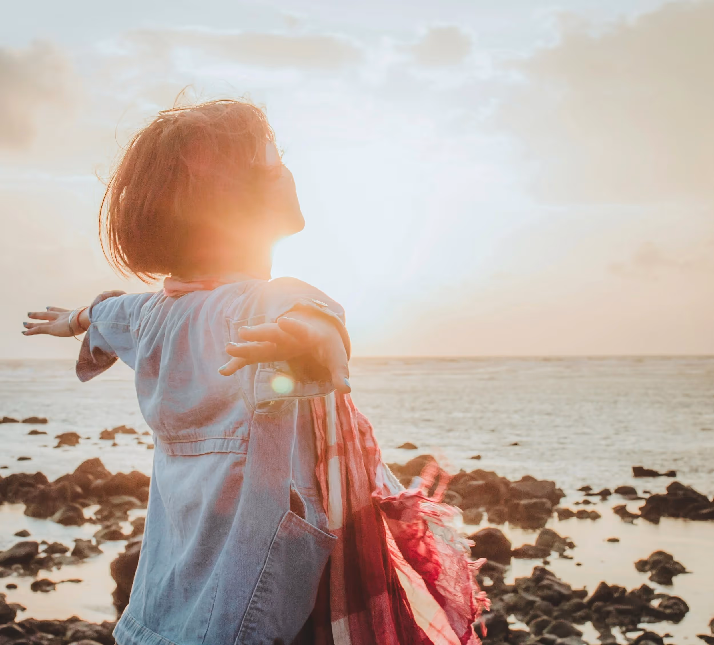 Person with short hair and a scarf extends arms wide facing the sea at sunset over rocky shore.
