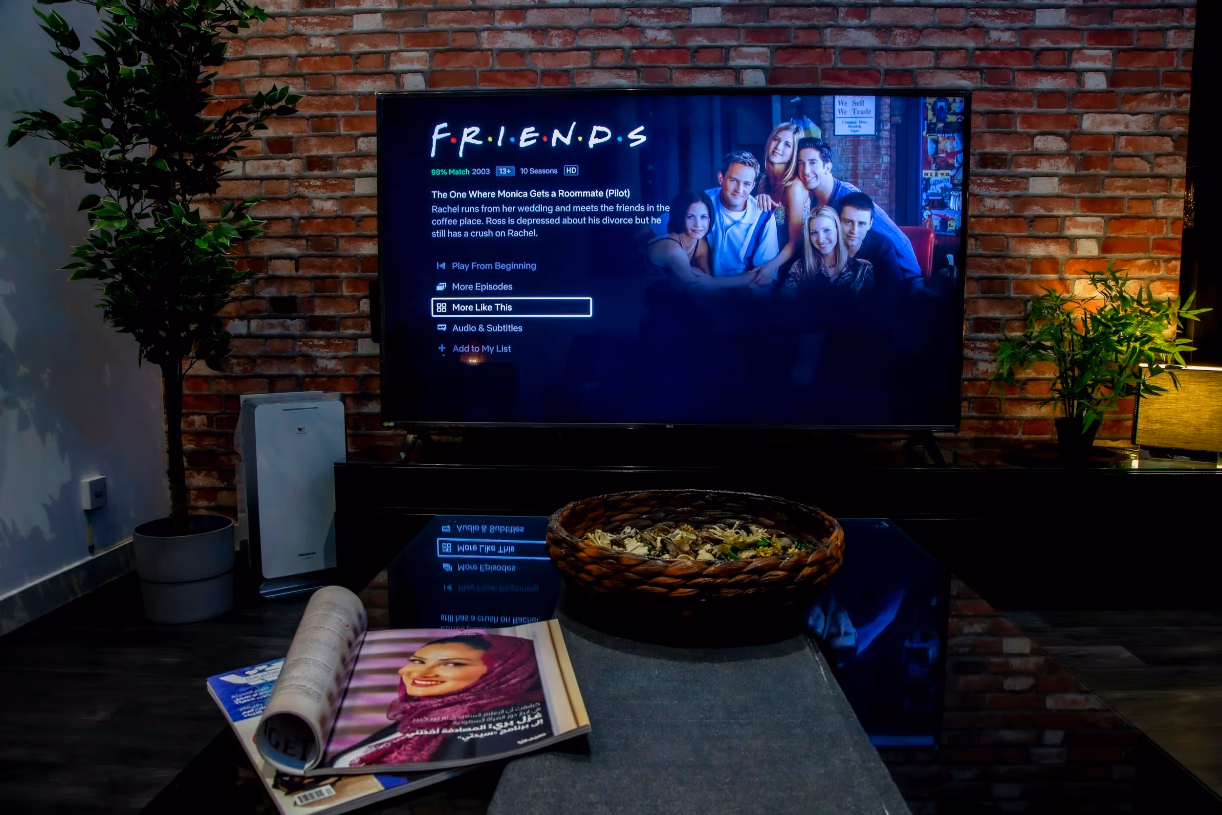 Living room with a TV showing the Friends series menu on a brick wall background, a coffee table with a magazine and a decorative bowl, and plants on both sides.