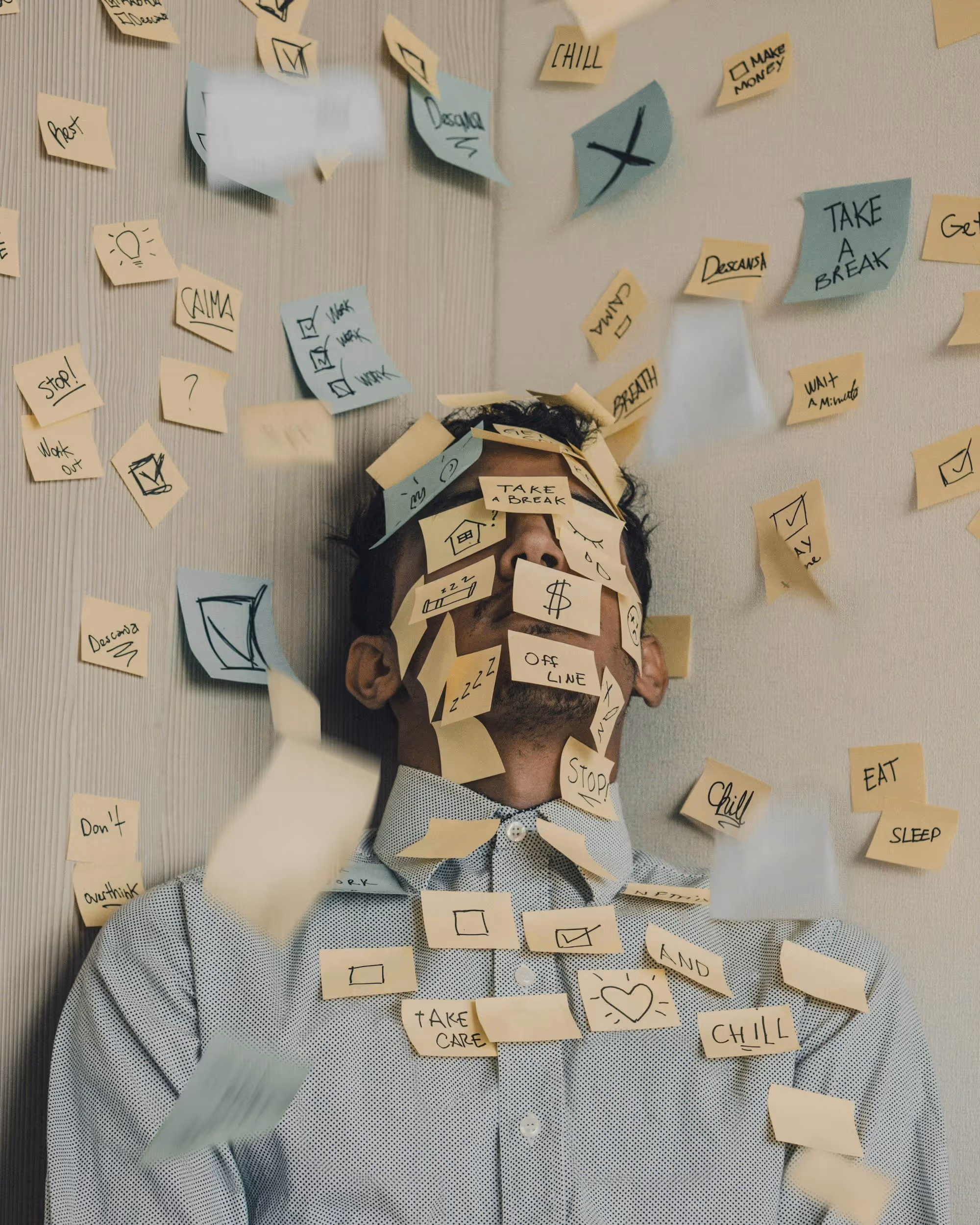 Man covered with numerous sticky notes featuring reminders like 'Take a break,' 'Chill,' 'Stop,' and 'Eat Sleep,' symbolizing overwhelm and stress.