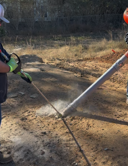 Two workers use a metal tool with a suction pipe on dry dirt in an outdoor worksite.