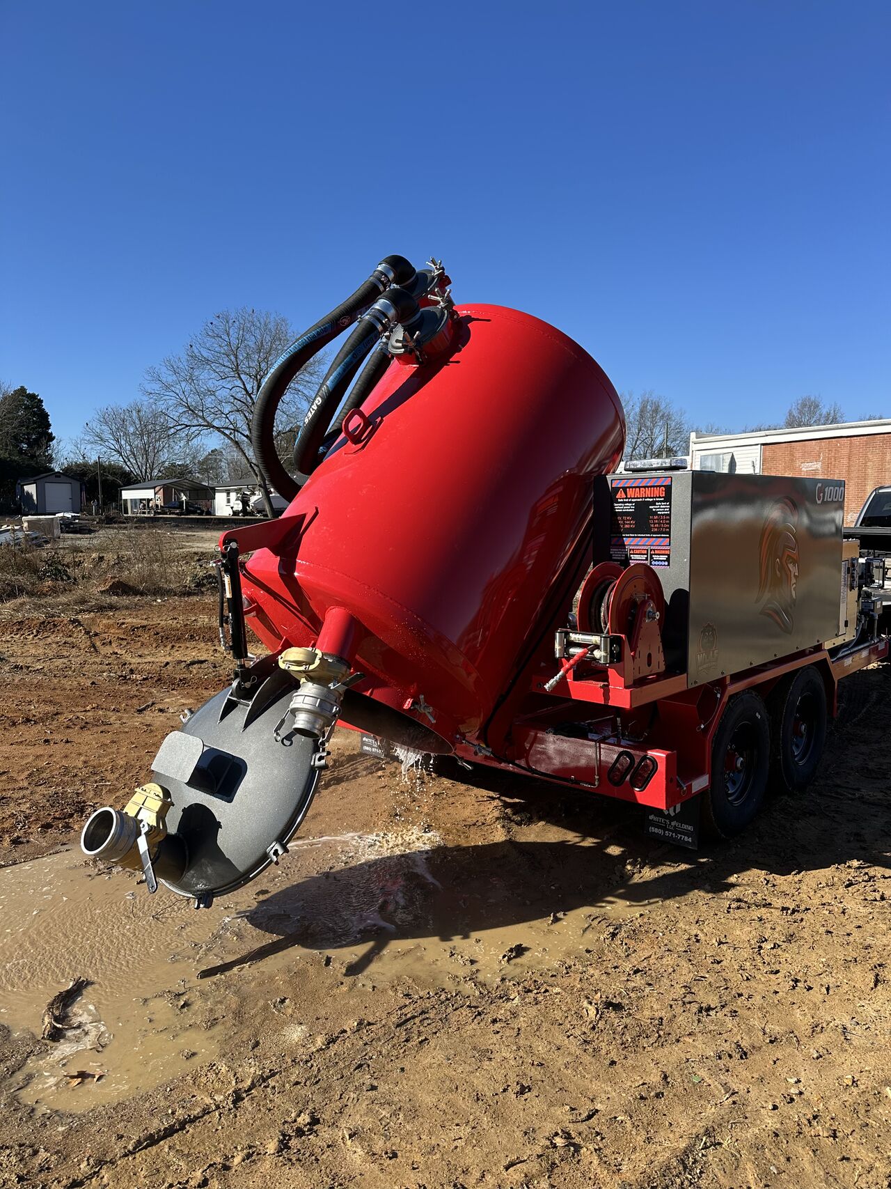 Red vacuum tank trailer tilted, draining muddy water on dirt ground under clear blue sky.