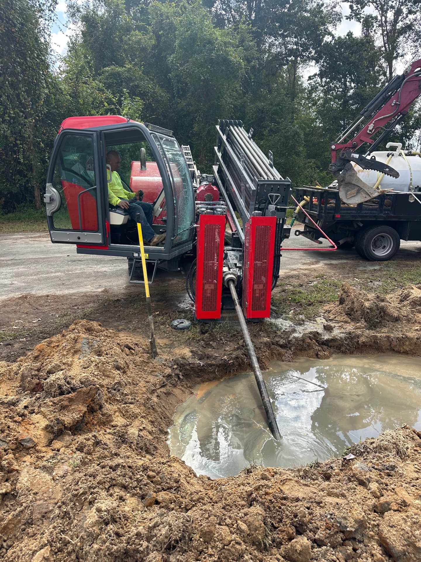 Worker operates horizontal drilling machine creating a muddy water hole near a wooded area.