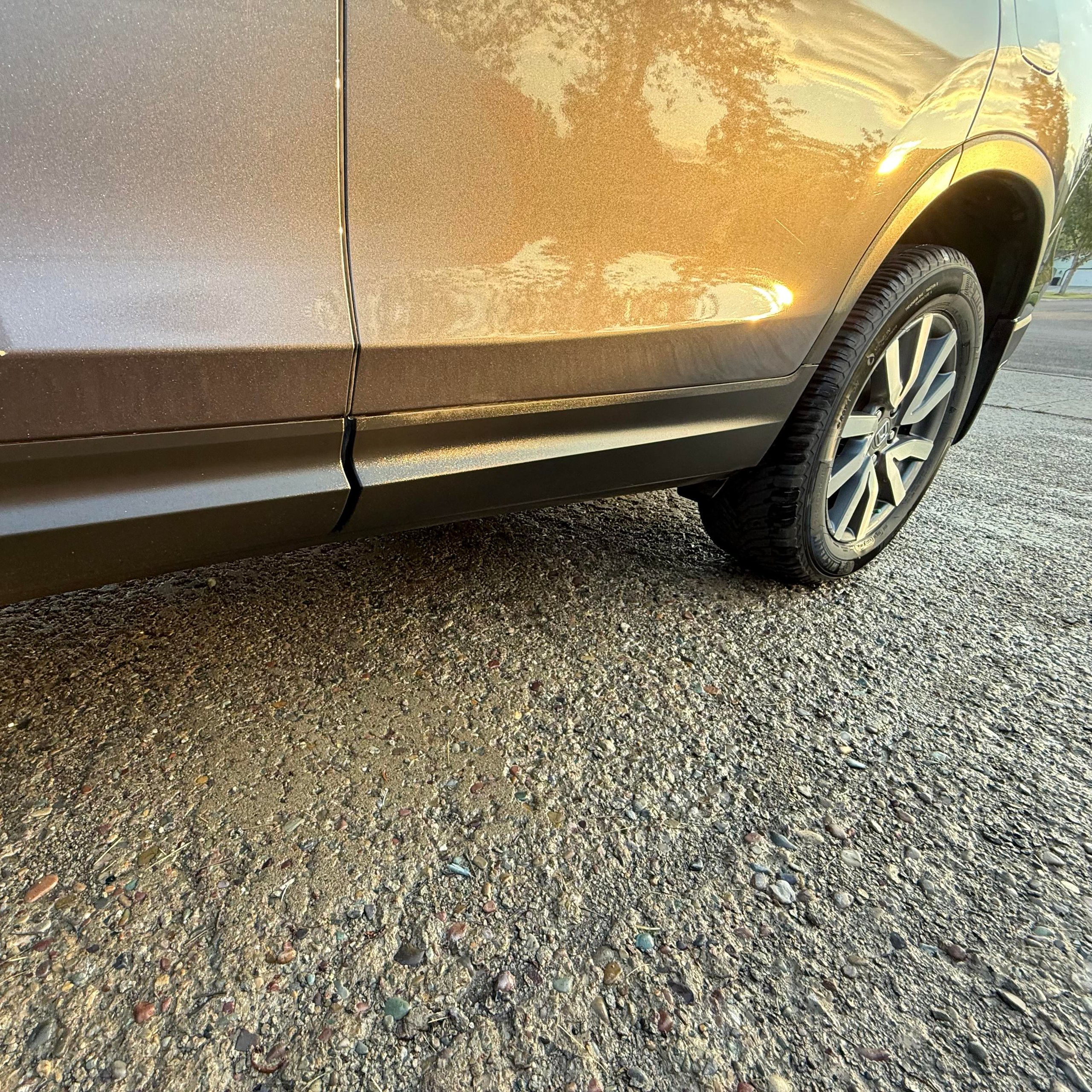 Close-up of a car's rear side door and rear wheel parked on a textured gravel surface with sunlight reflecting on the metallic paint.