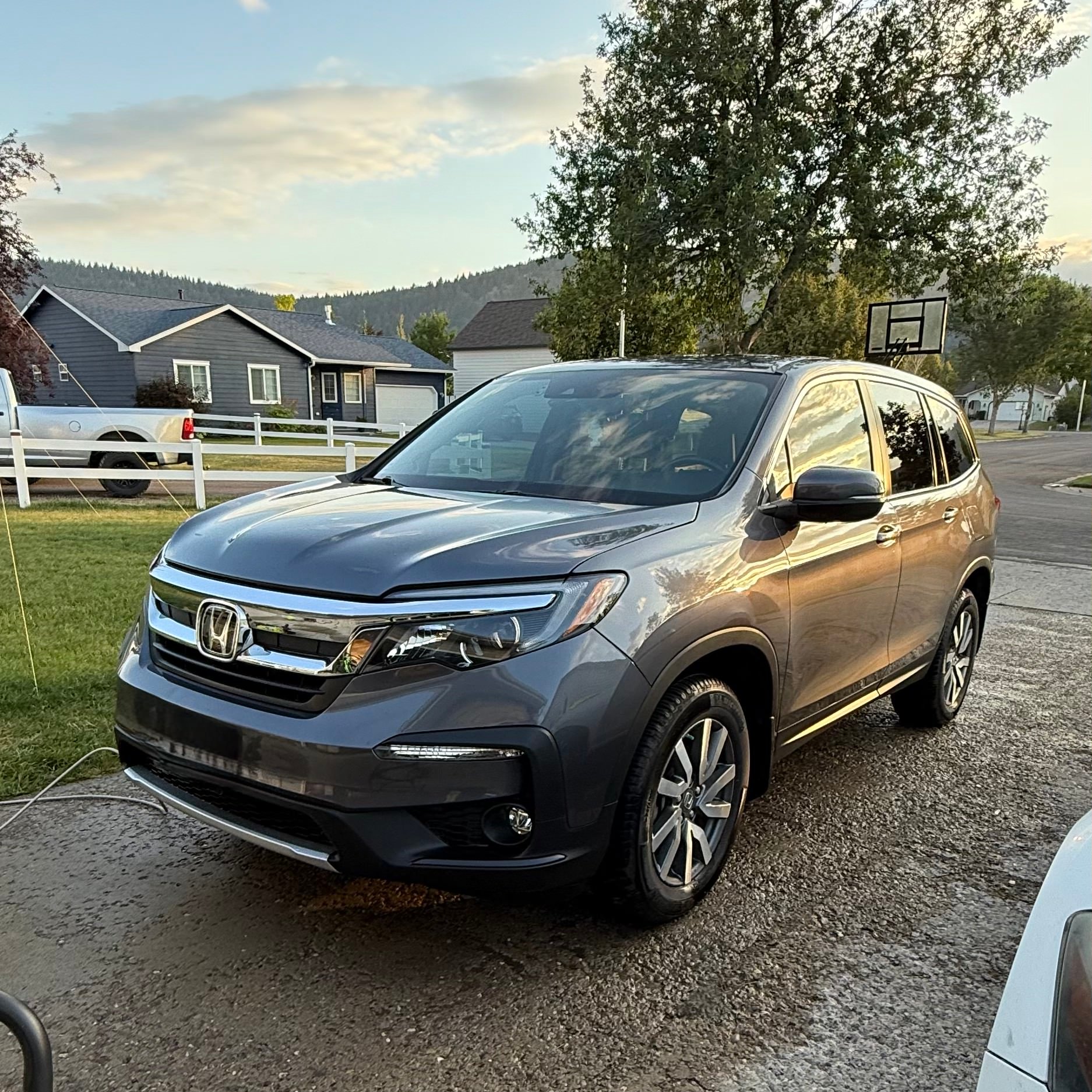 Gray Honda SUV parked on a driveway with houses, trees, and a basketball hoop in the background during sunset.