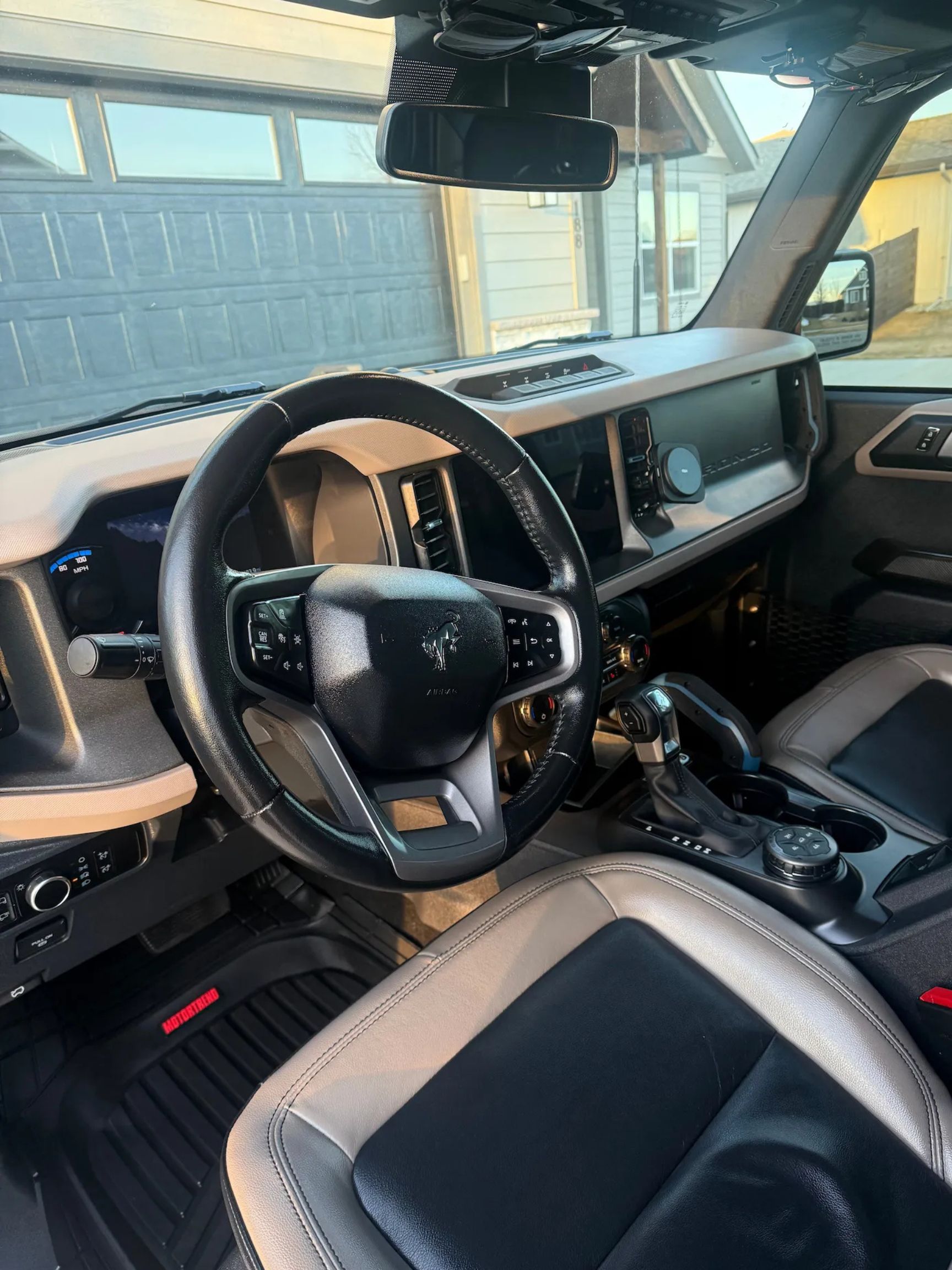Interior of a modern vehicle showing steering wheel with Ford Bronco logo, dashboard, gear shift, and two-tone leather front seat.
