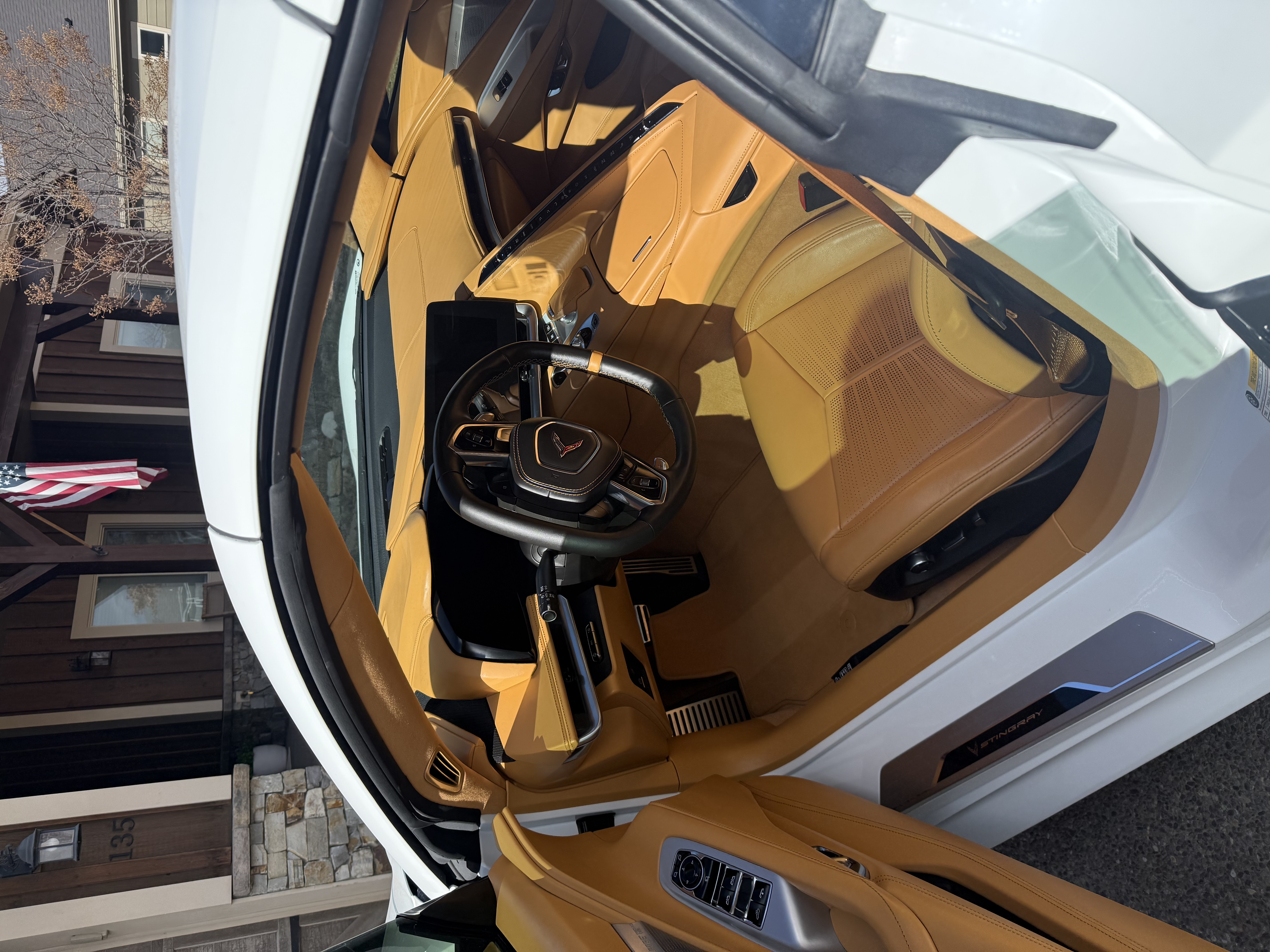 Interior of a white Corvette Stingray showing a tan leather driver seat, dashboard, and steering wheel with digital display.