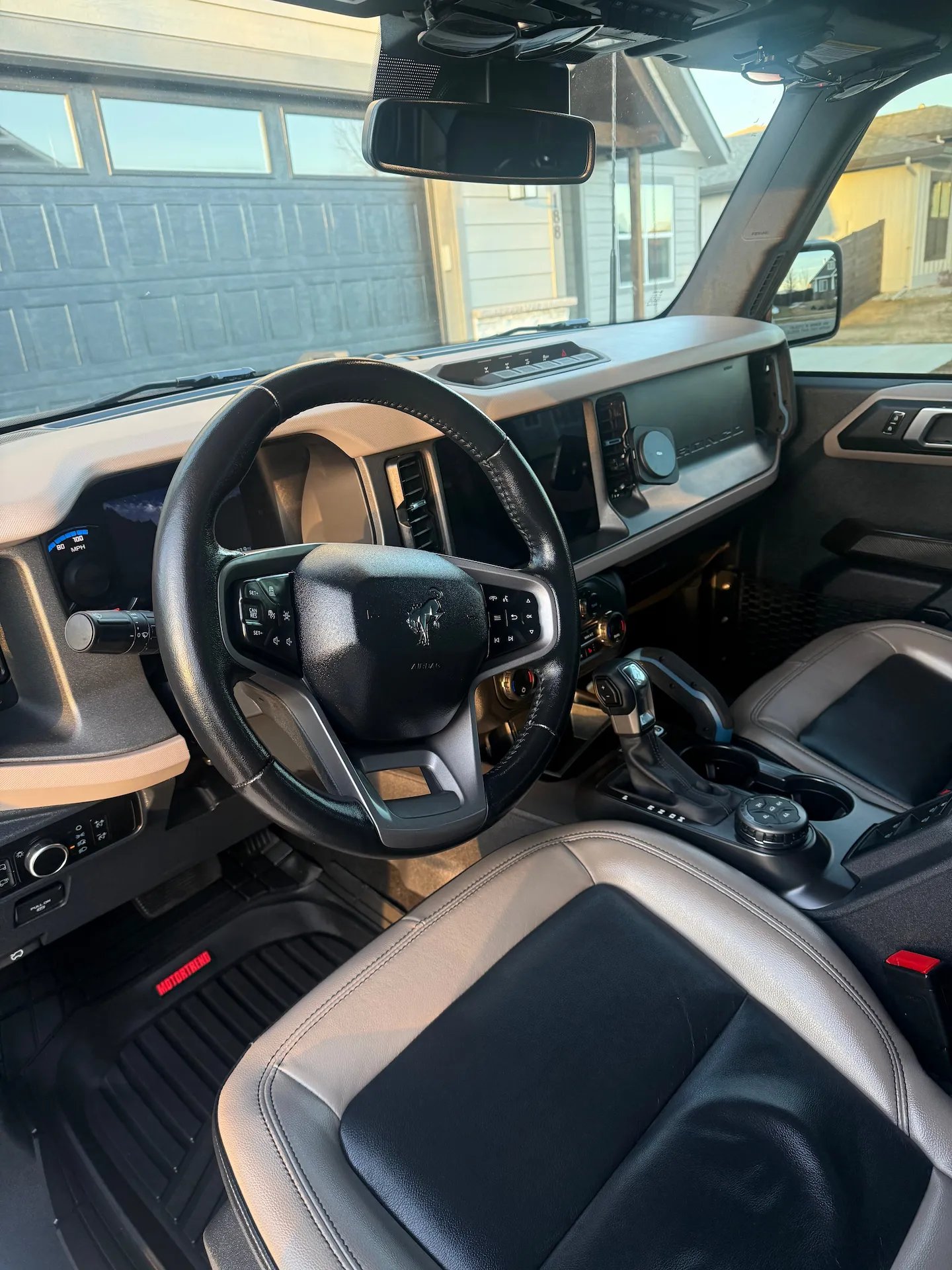 Interior view of a vehicle showing a black and gray leather driver's seat, steering wheel with controls, gear shift, and dashboard with digital display.