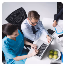Two healthcare professionals working together on a laptop at a desk with documents, tablet, and green apples.
