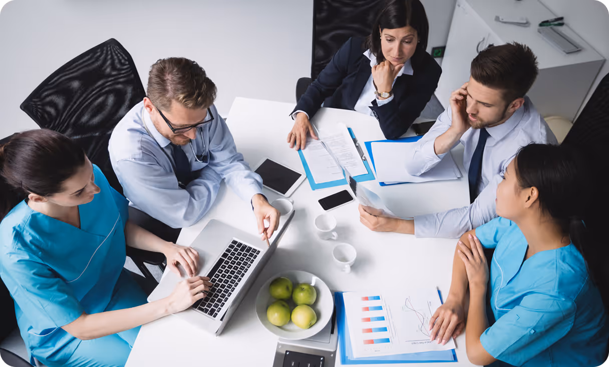 Group of five medical professionals and businesspeople sitting around a table reviewing documents and working on a laptop.