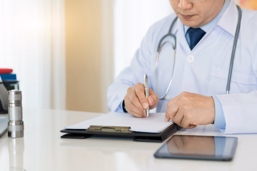 Doctor in white coat with stethoscope writing on clipboard at desk with tablet and flashlight.