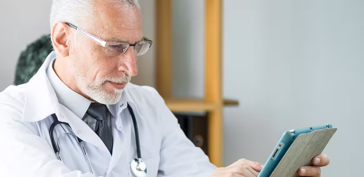 Senior male doctor with glasses and stethoscope using a tablet in a medical office.