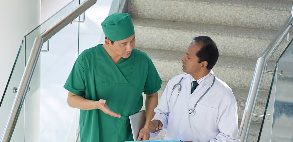 Two male doctors in scrubs and white coat discussing medical documents on stairs.
