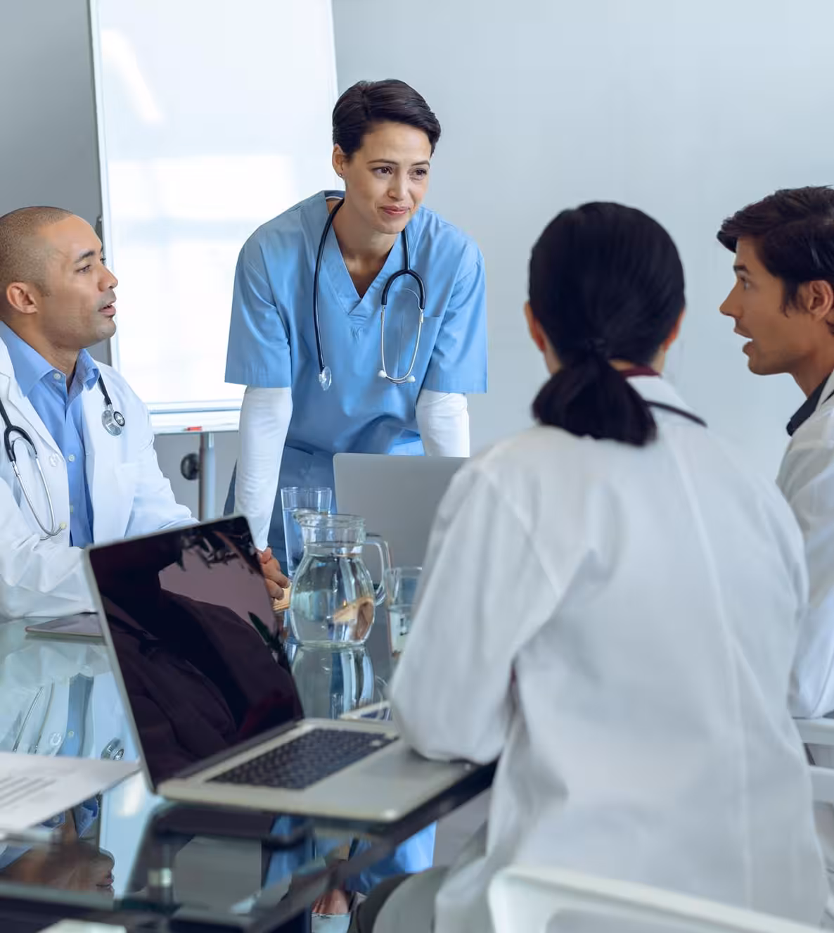 Doctors and a nurse in scrubs discussing at a glass table with laptop and water jug in a meeting room.