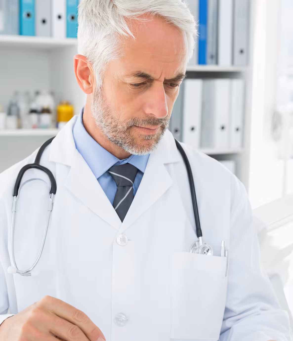 Mature male doctor with gray hair and stethoscope, focused on writing or reading in clinic.