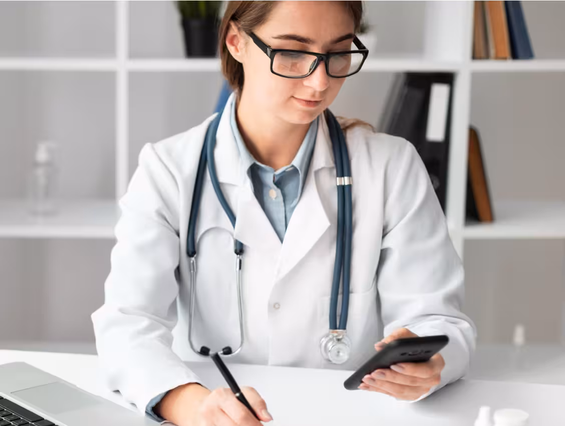 Female doctor in white coat with stethoscope using phone and writing notes at desk.