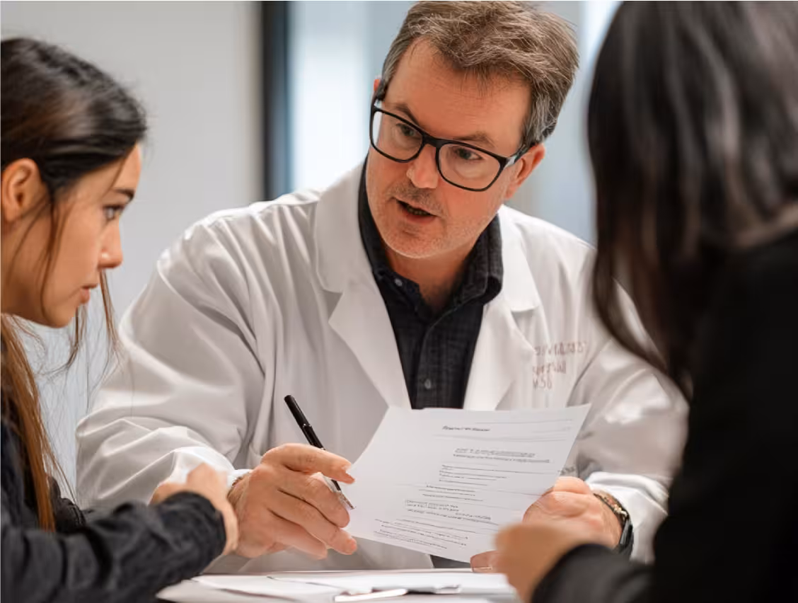A male doctor in a white coat explains a document to two women across a table in a consultation.