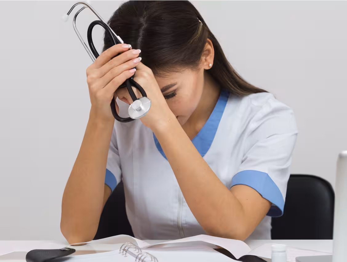 Stressed medical professional holding a stethoscope to her head at a desk with open notebook.