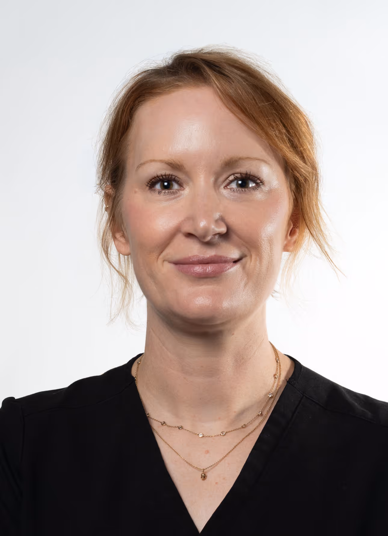 Portrait of a smiling woman with red hair wearing a black top and gold necklaces.