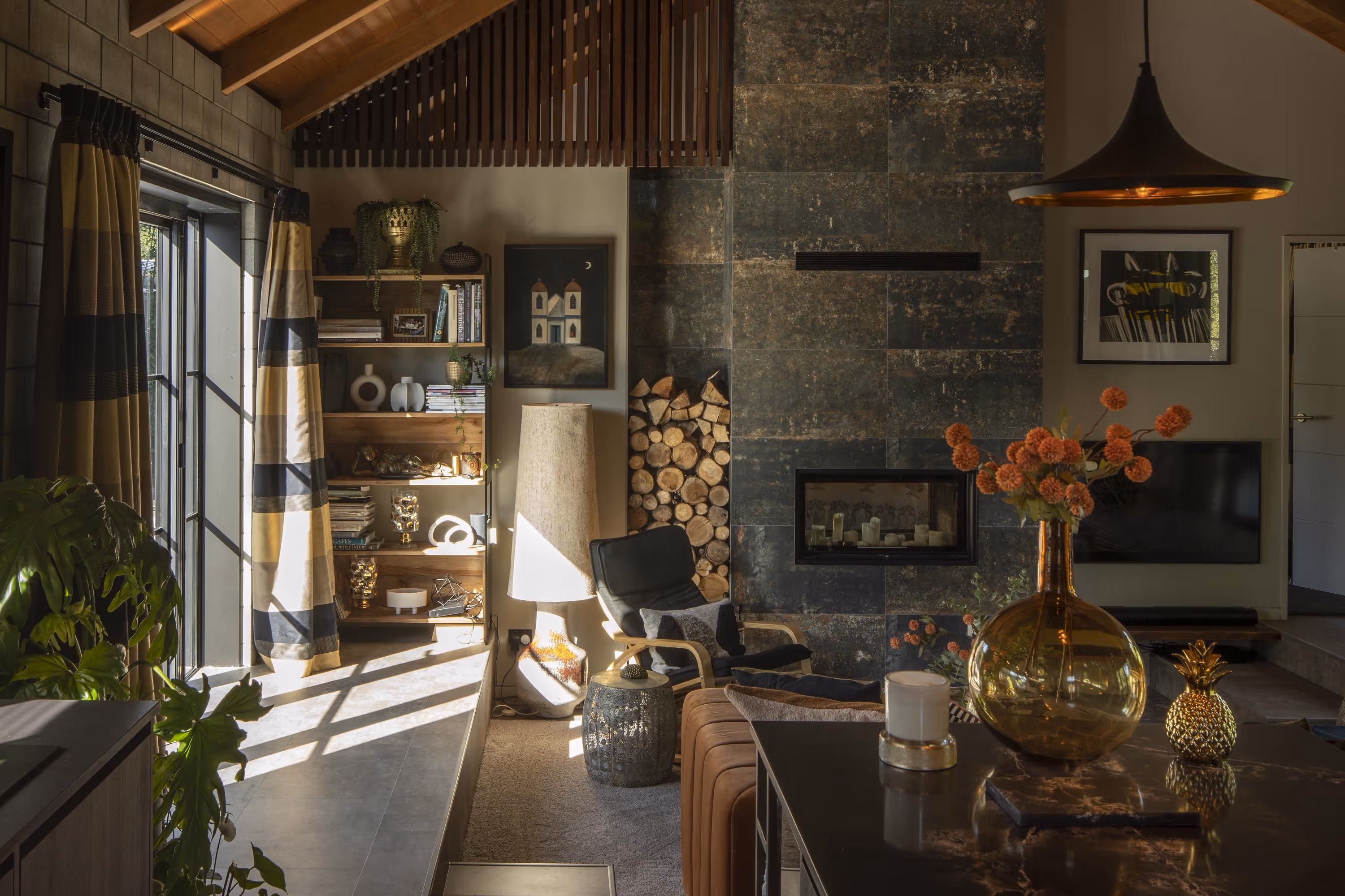 Cozy living room with sunlight streaming through large windows, featuring a black lounge chair, stacked firewood, a dark stone fireplace, shelves with decor, and a table with a vase of orange flowers.