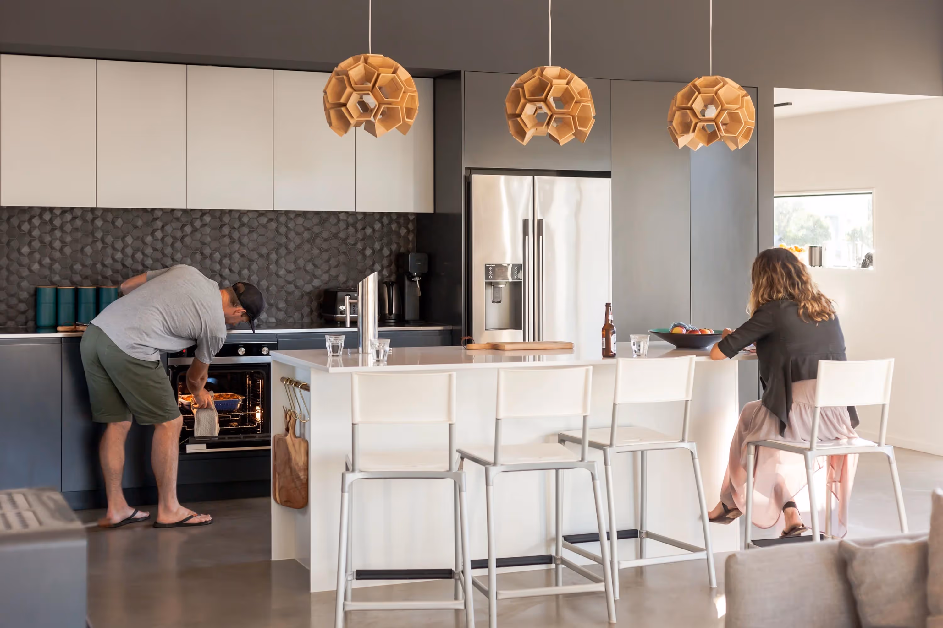 Modern kitchen at the Saunders Residence in Greytown with white island, wooden pendant lights, and two people using the space.
