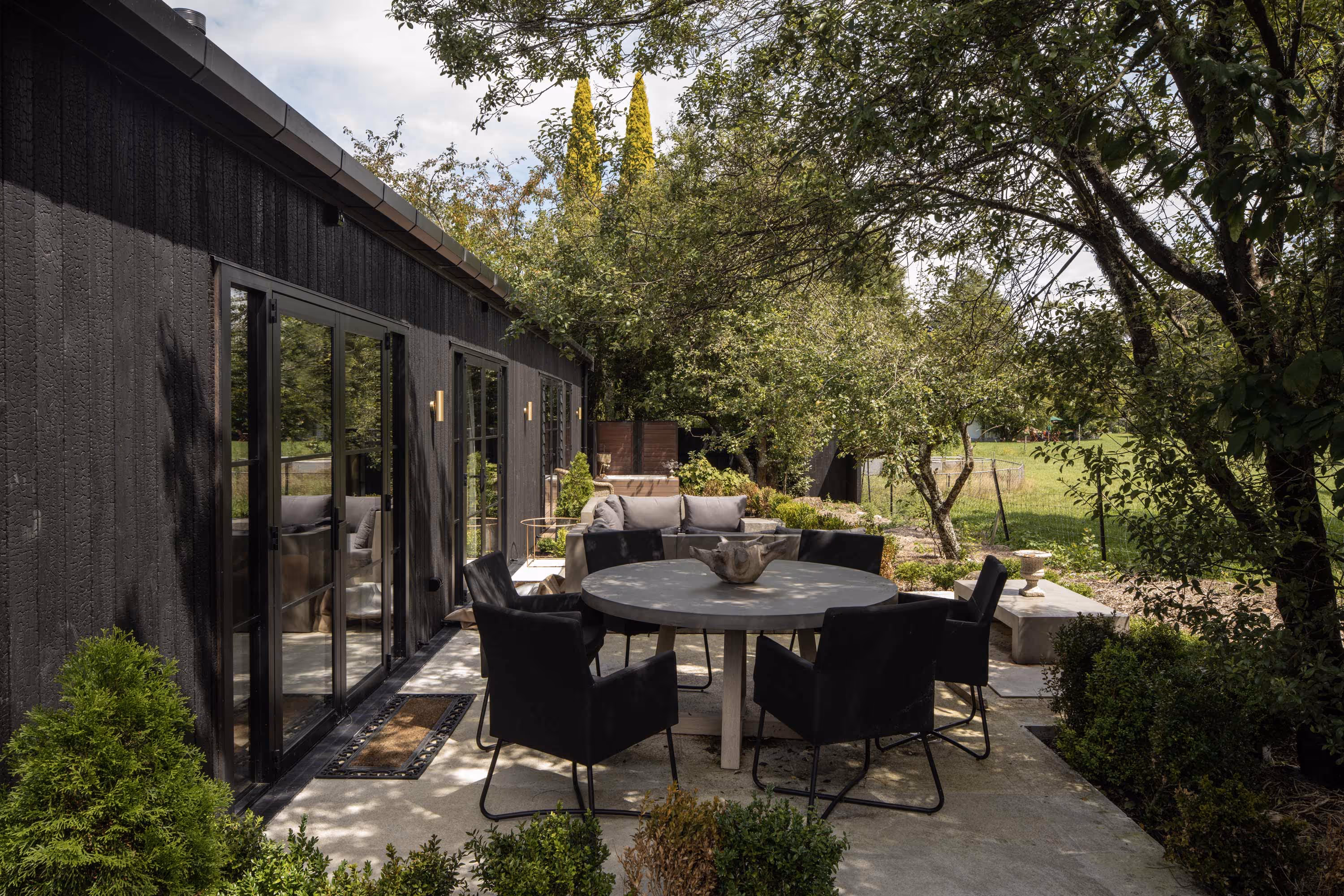 Outdoor patio at the Smith House in Greytown with round table, black chairs, gray sofa, and black-framed glass doors on timber-paneled house.