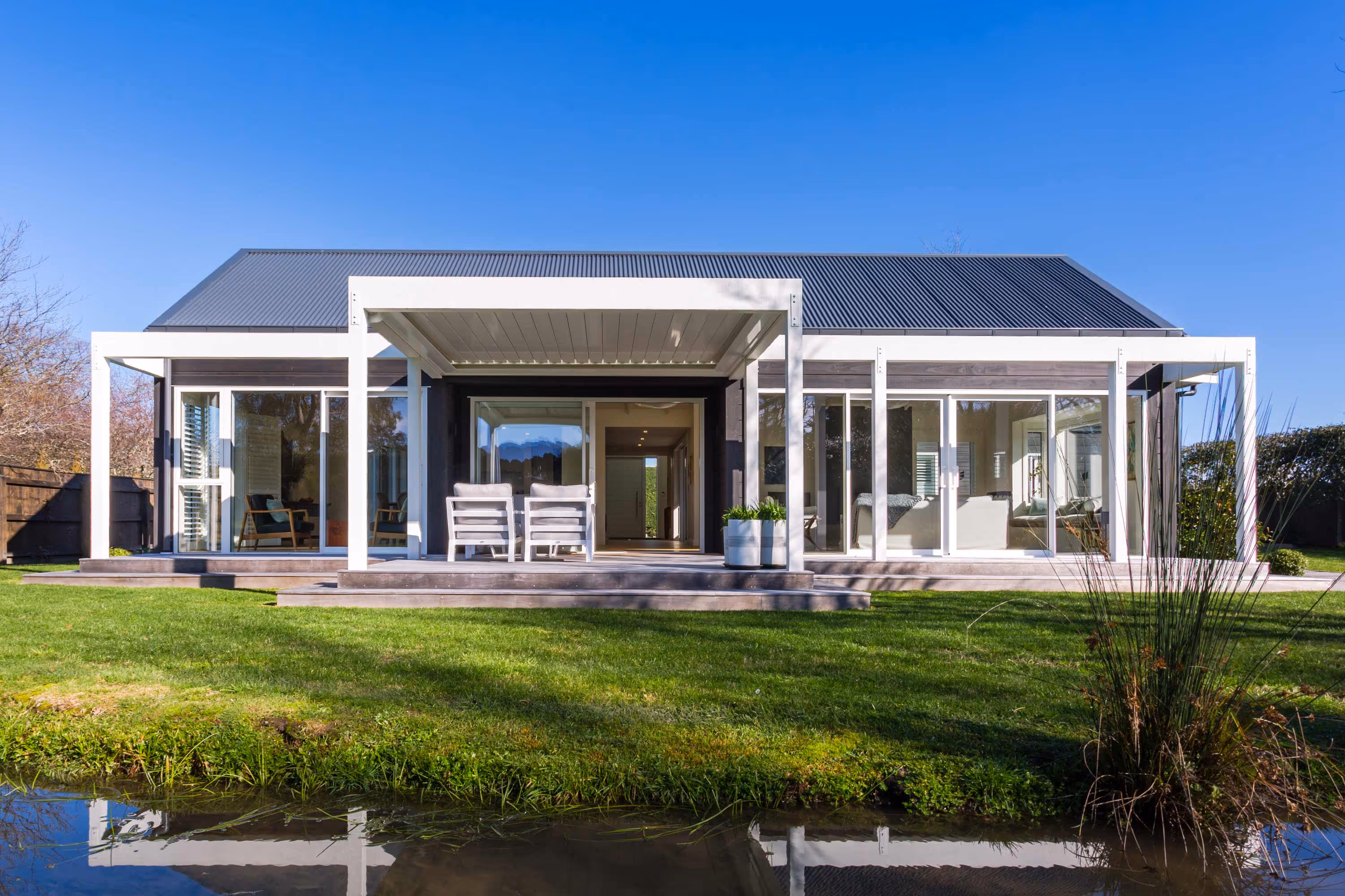 Kemp House in Greytown, dark timber home with large glass windows, patio, green lawn, and reflection in pond.