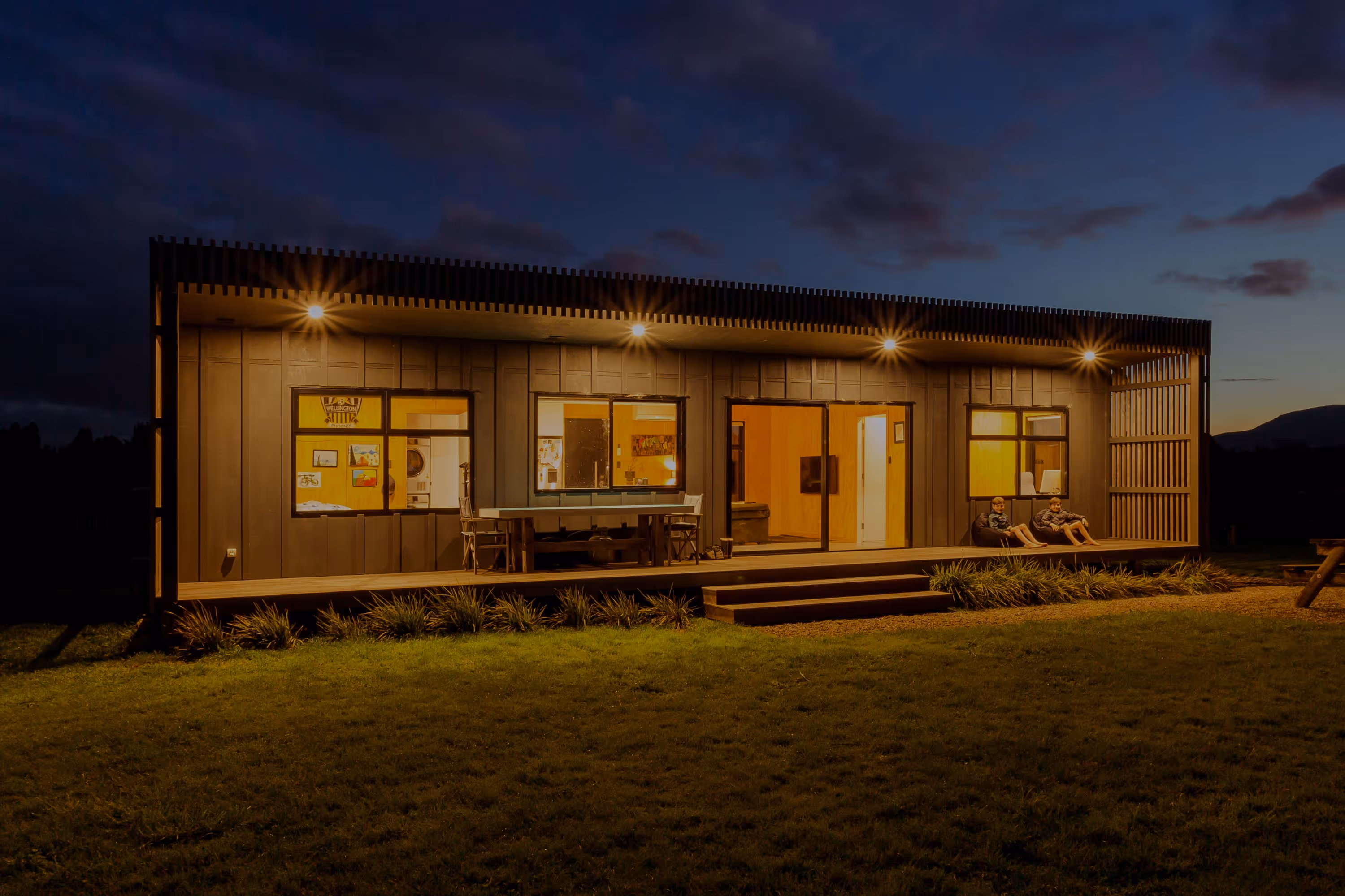 Modern house with illuminated porch at dusk, two children sitting on the right side of the deck.