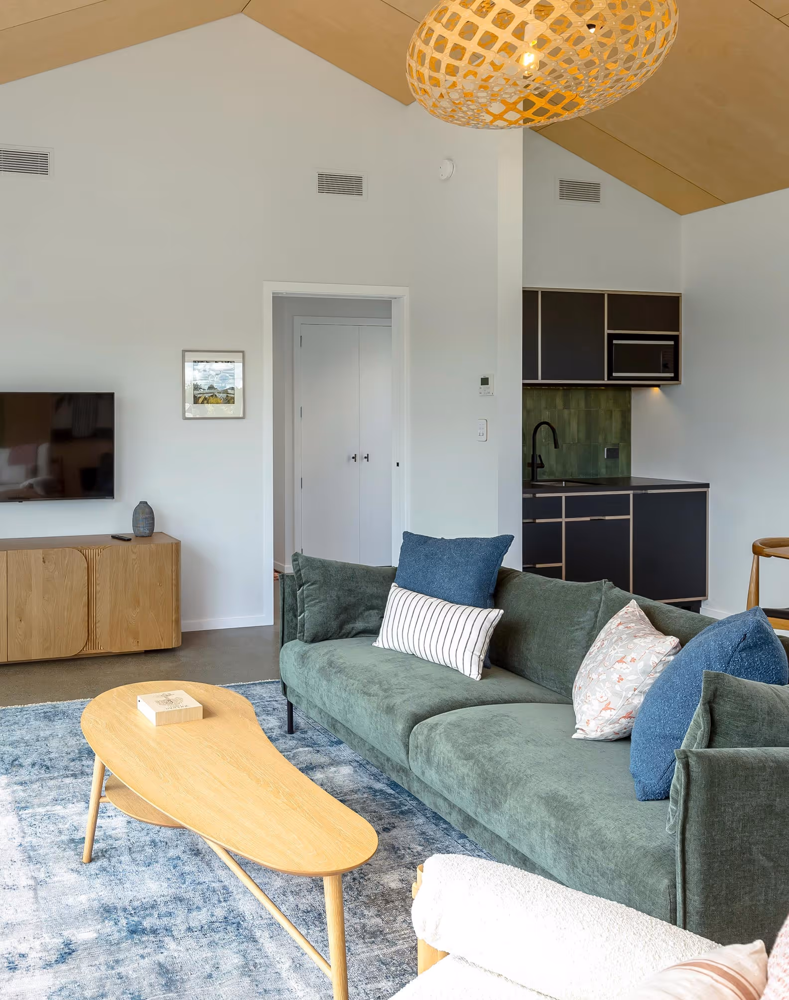 Modern living room at Fisher & Clarke house, Martinborough, with green sofa, patterned pillows, light timber coffee table, and kitchenette with dark cabinets.