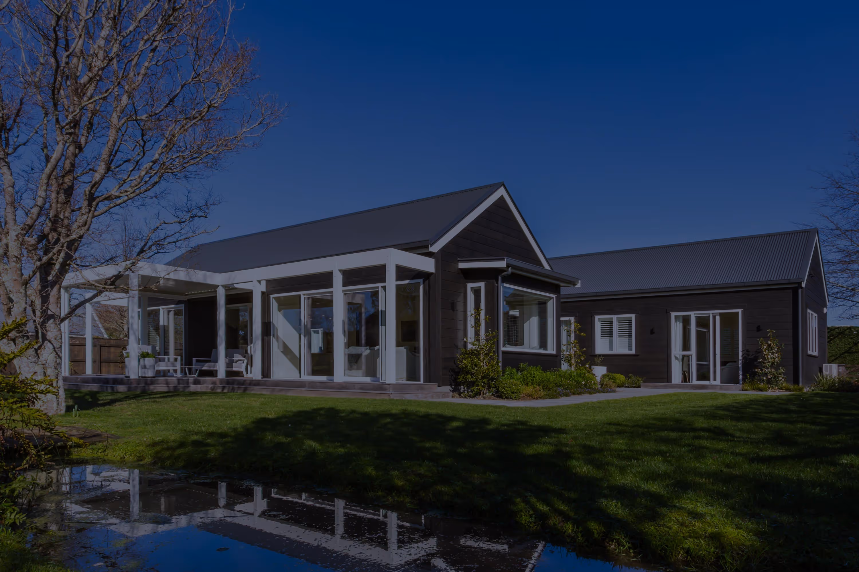 Kemp House in Greytown, dark timber home with large glass windows, patio, green lawn, and reflection in pond.