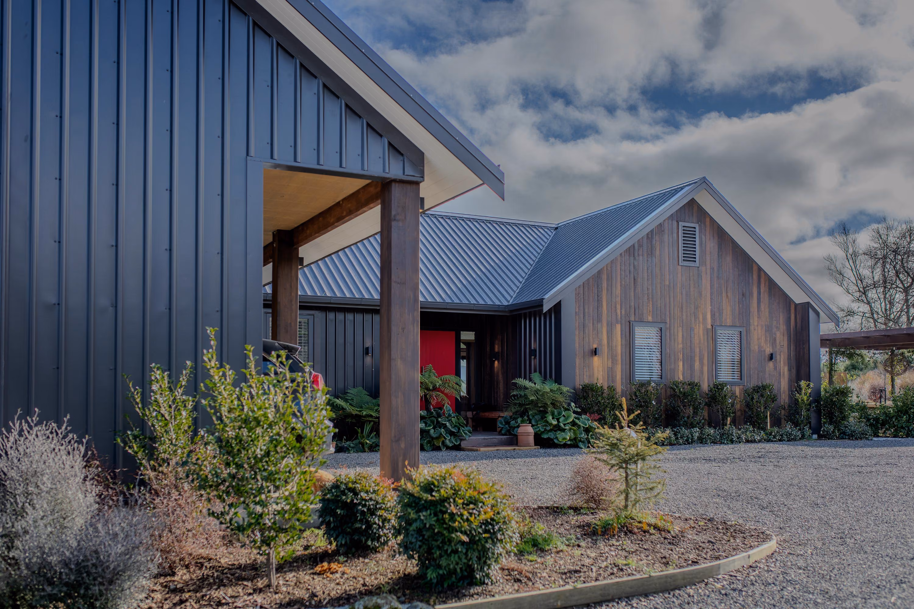Front exterior of the home with dark vertical cladding, timber entrance porch, red door and landscaped gravel driveway