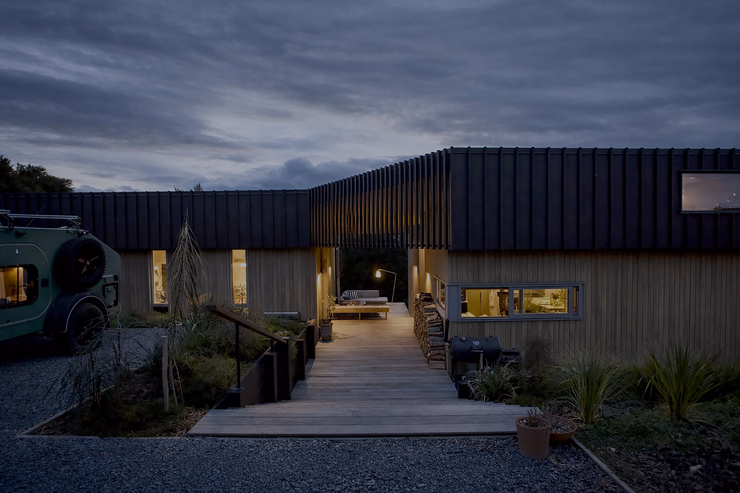 Modern house exterior at dusk with warm interior lights, wooden walkway entrance, stacked firewood, and green plants, under a cloudy sky.
