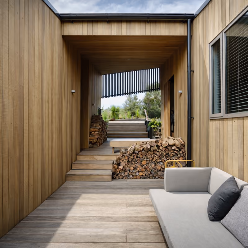 Modern outdoor patio area with wooden flooring and walls, a light gray cushioned sofa with dark pillows on the right, stacked firewood, steps leading to a raised deck with plants and outdoor furniture under a partly cloudy sky.