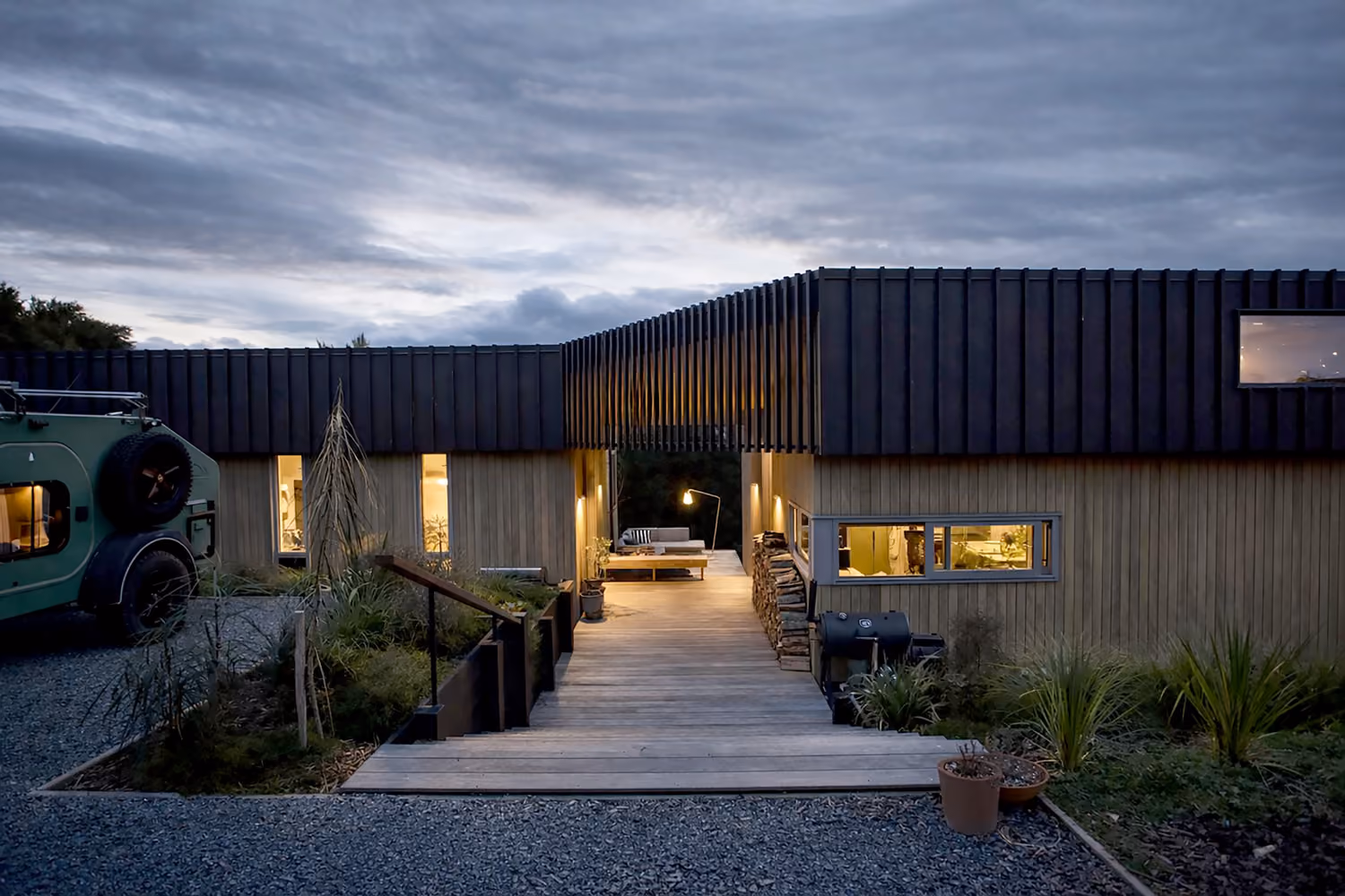 Modern house exterior at dusk with warm interior lights, wooden walkway entrance, stacked firewood, and green plants, under a cloudy sky.