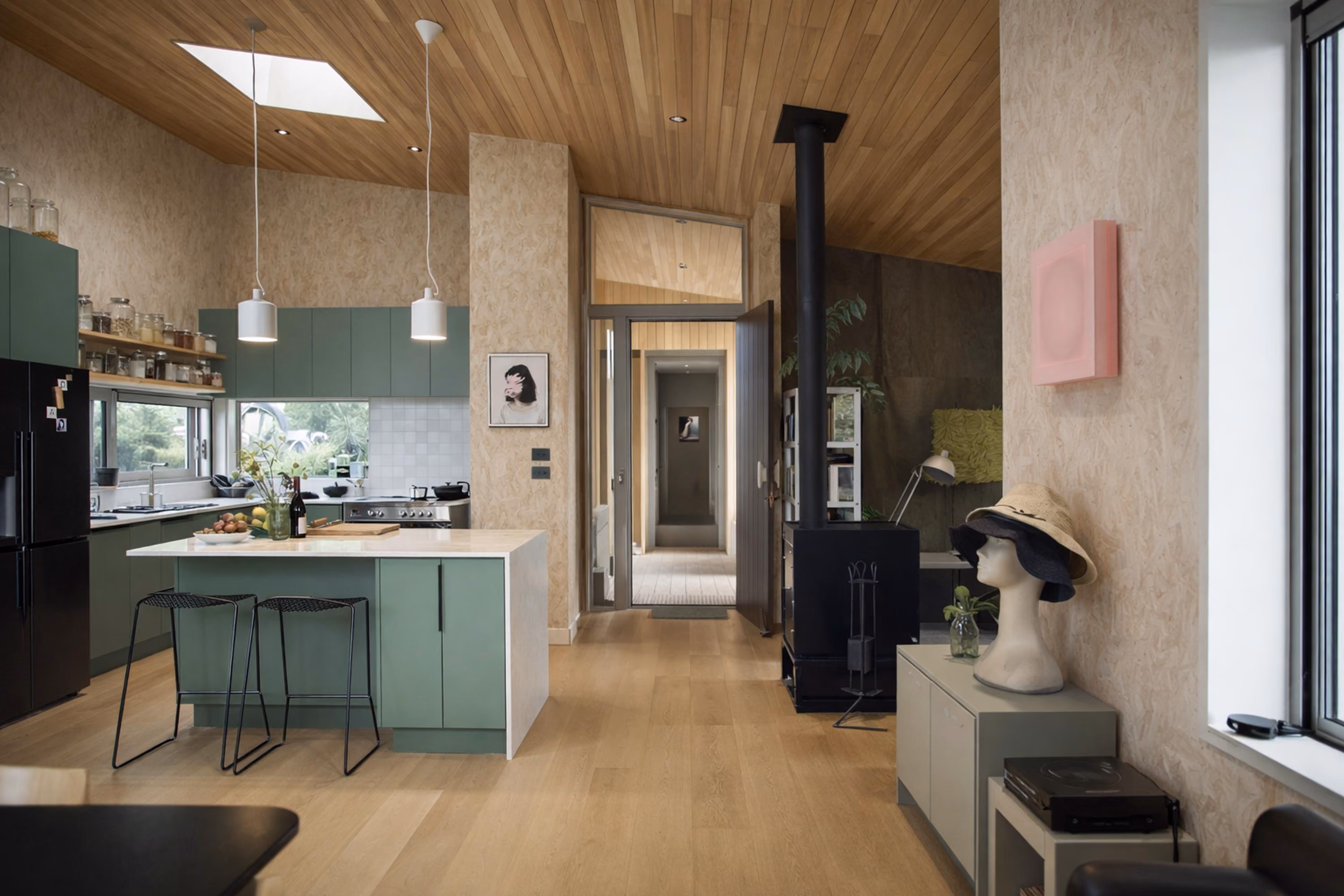 Modern open kitchen and living area with wood ceiling and floor, green cabinetry, black refrigerator, white island with two stools, wood-burning stove, and natural light from windows and skylight.