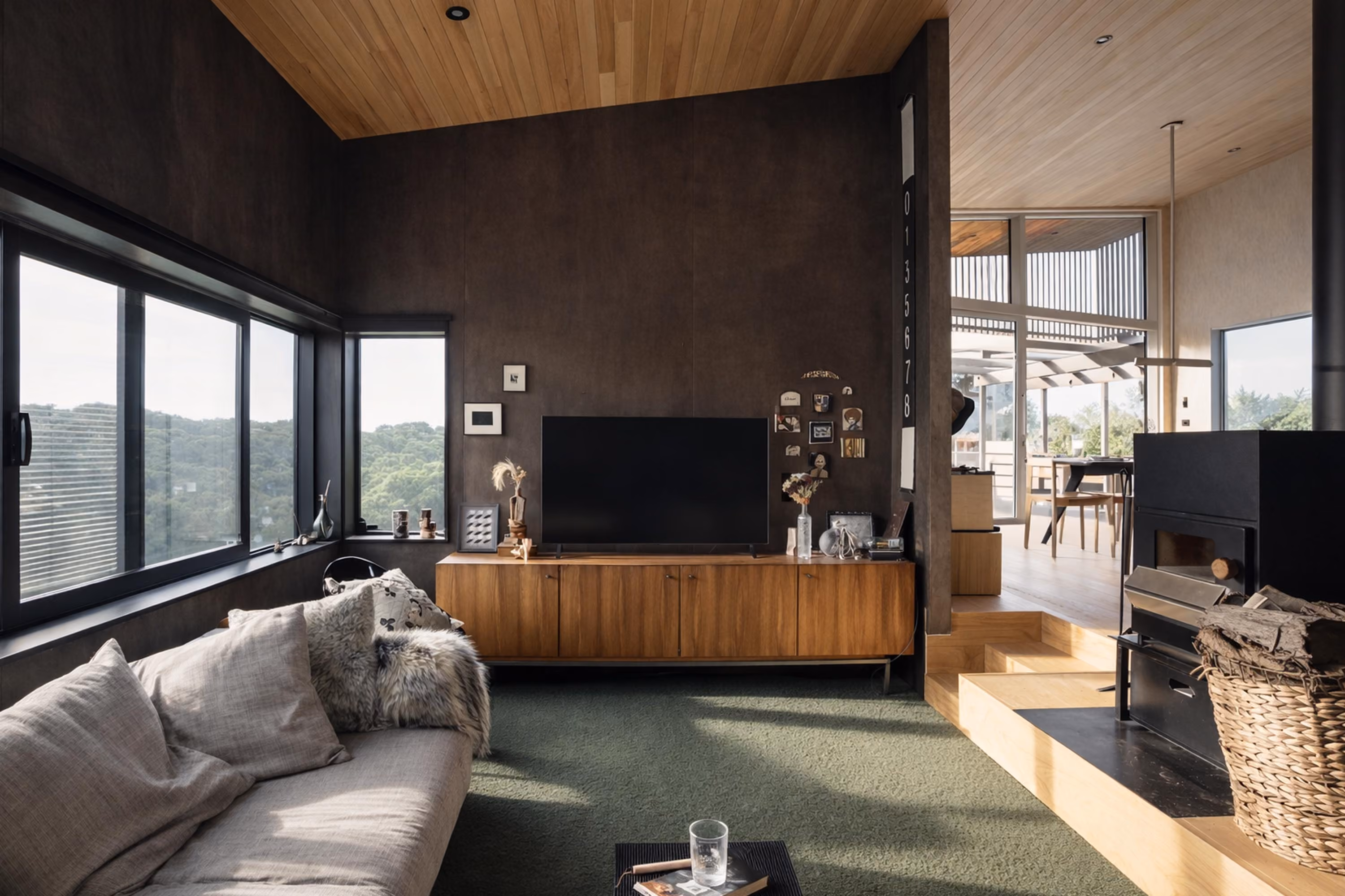 Modern living room with large windows overlooking greenery, a beige sofa with cushions and a fur throw, a wooden TV cabinet with a flat-screen TV and decorative items, a carpeted floor, and a nearby black fireplace with a basket of firewood next to a step leading to a dining area.