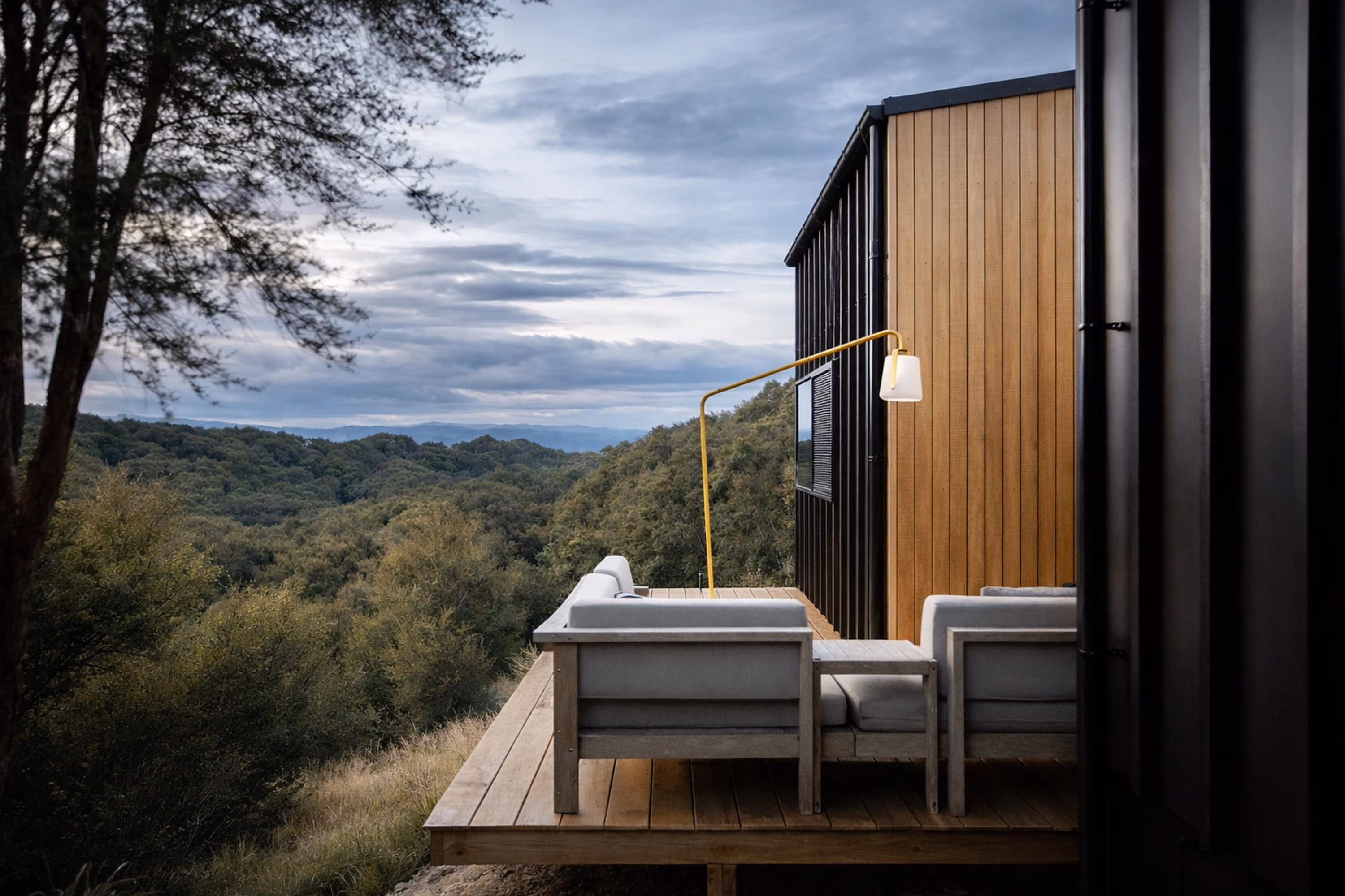 Wooden deck attached to a modern house with white cushioned outdoor chairs and a yellow floor lamp, overlooking a forested landscape under a cloudy sky.