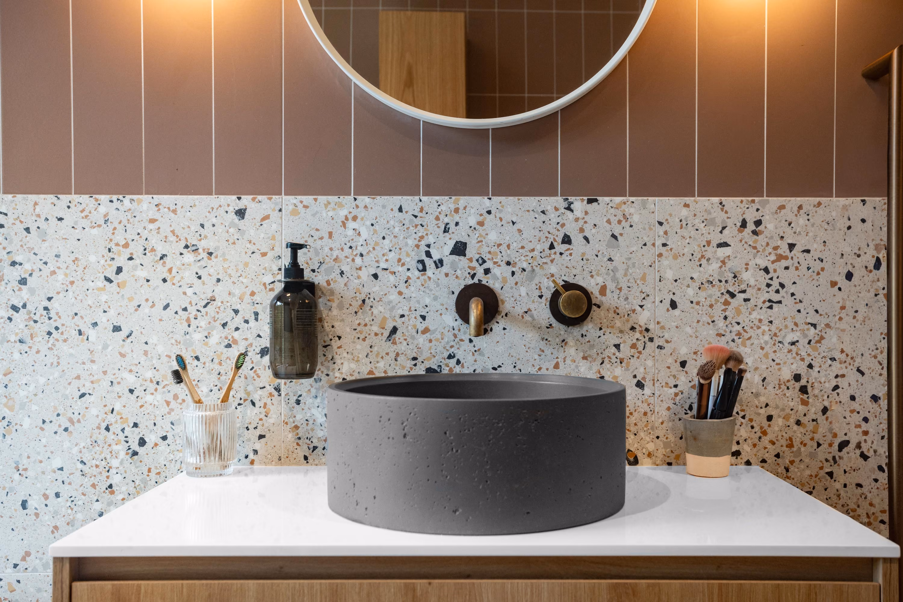 Modern bathroom sink with a round, dark gray basin on a white countertop with wood cabinetry below, terrazzo wall tiles behind, wall-mounted brass faucet and soap dispenser, and toothbrush and makeup brush holders on either side.