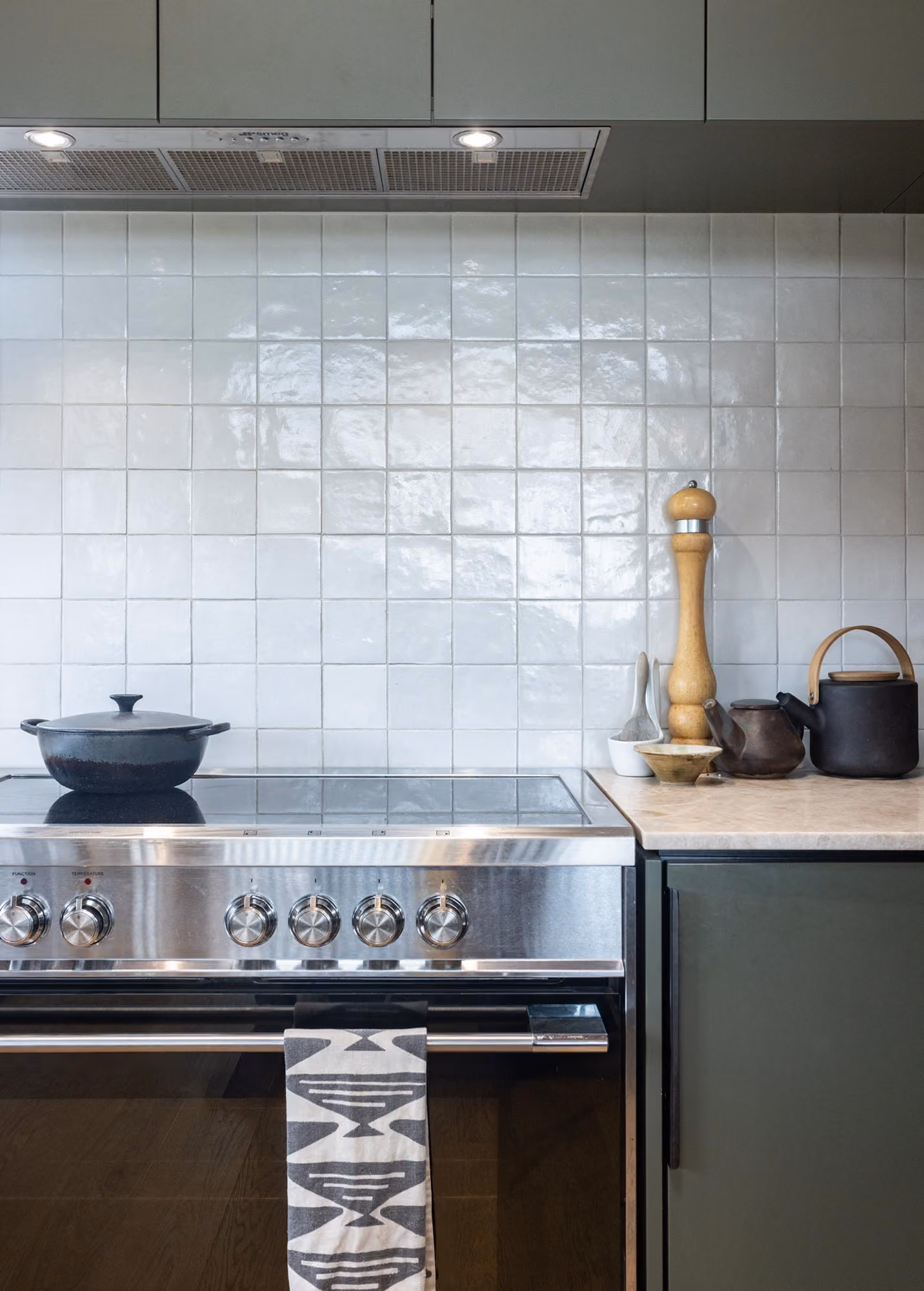 Modern kitchen stove with stainless steel oven and black cooktop, a black pot on the stove, and a countertop with a wooden pepper grinder, ceramic containers, and a black teapot with a wooden handle against a white tiled backsplash.