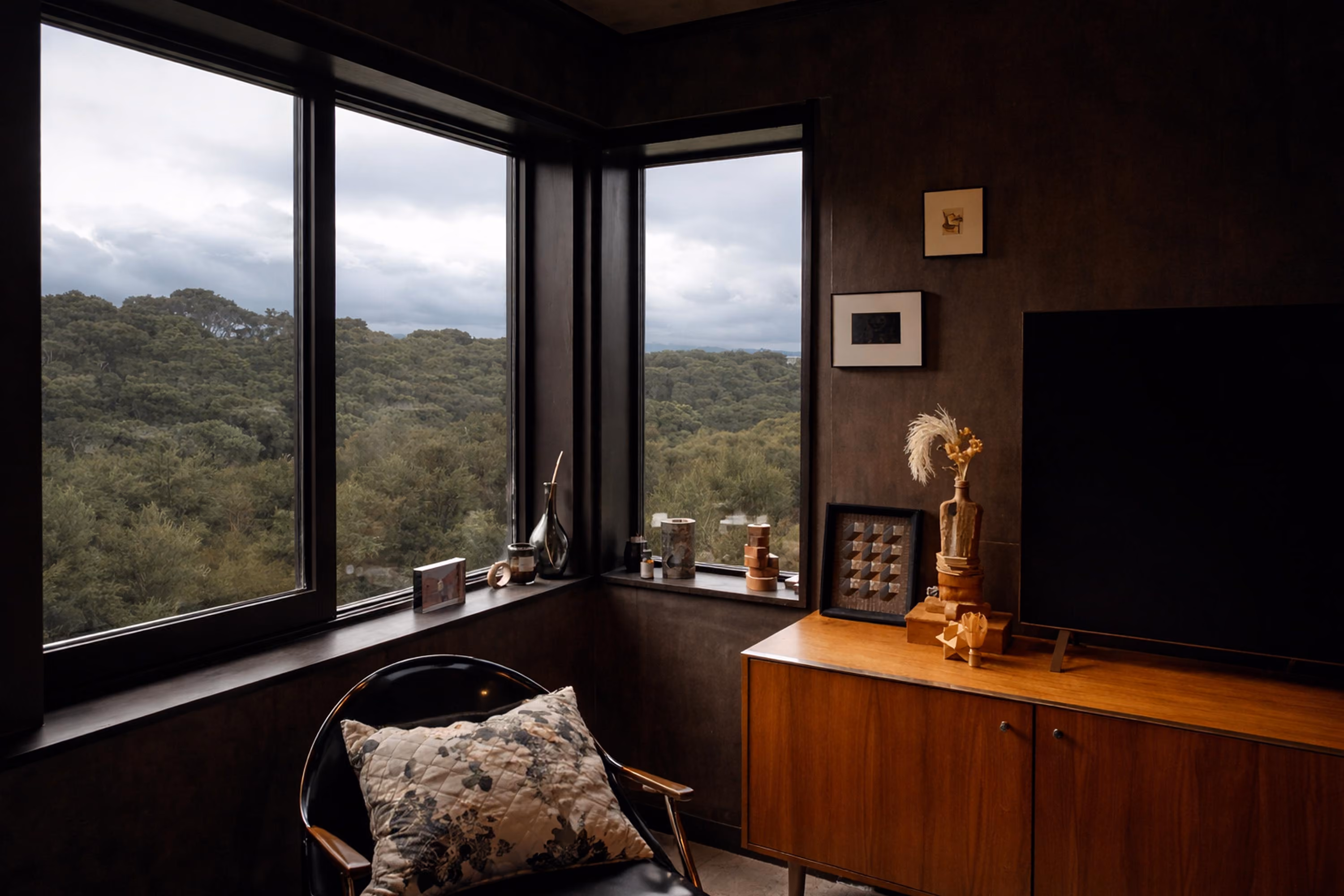 Cozy corner of a room with large windows showing a forest view under a cloudy sky, a black chair with a patterned pillow, and a wooden cabinet with decorative items and a TV.