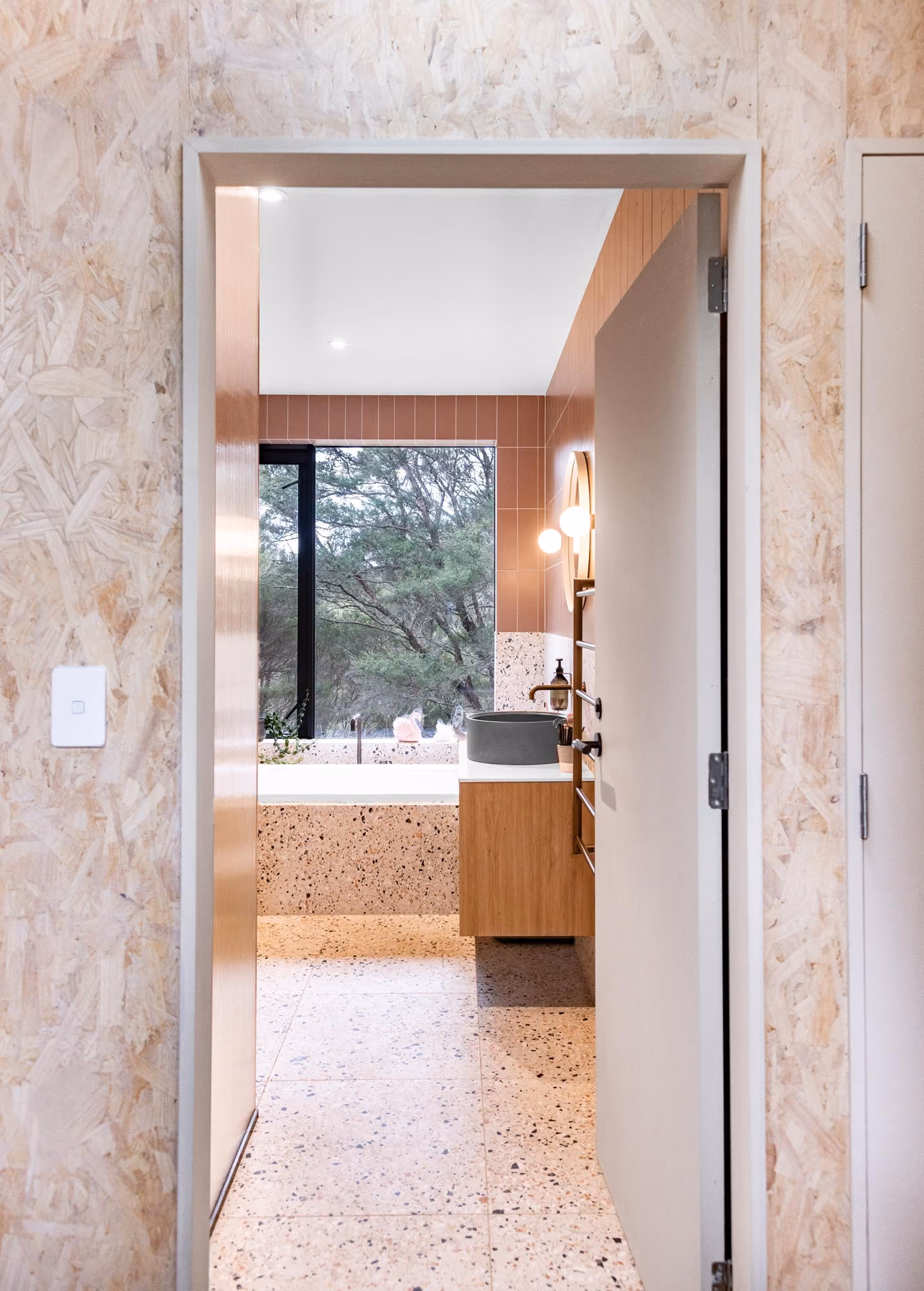 View through a doorway into a modern bathroom with terrazzo floor and bathtub surround, a wooden vanity with a gray vessel sink, wall-mounted faucet, round mirror, and a large window showing trees outside.
