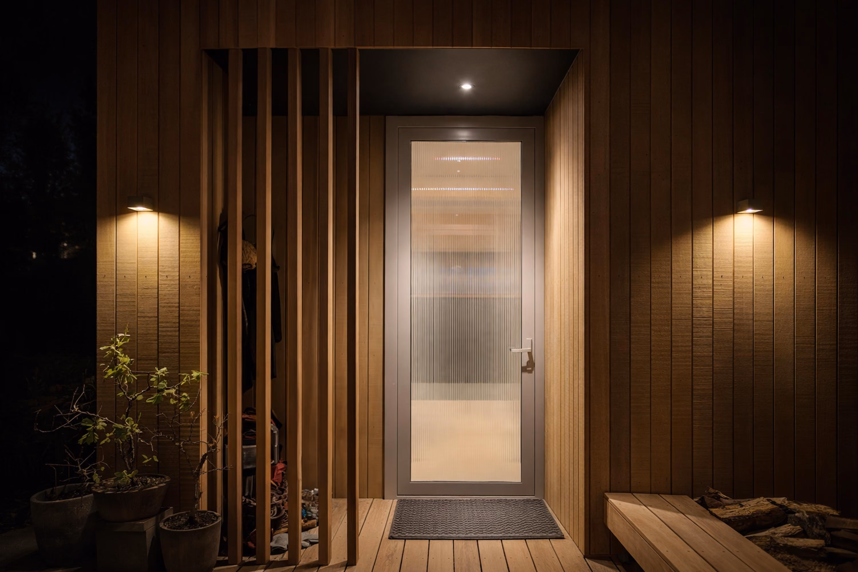 Modern wooden front entrance at night with vertical wood slats, a frosted glass door, wall-mounted lights on either side, potted plants on the left, and a wooden bench with firewood on the right.
