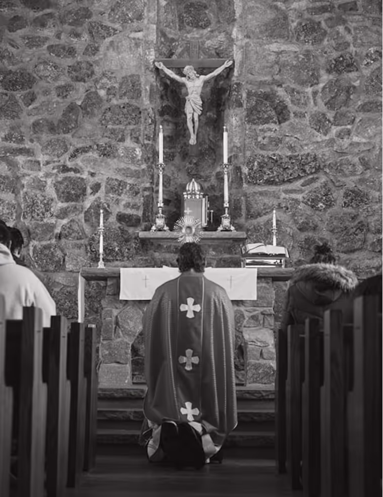 Priest kneeling in prayer facing an altar with a crucifix and tabernacle in a Catholic church.